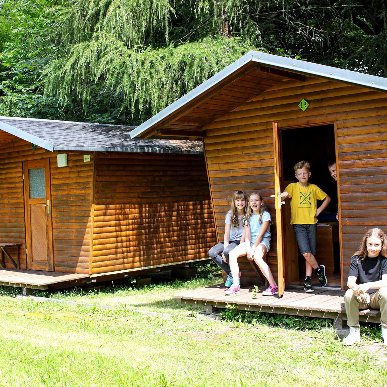 Zwei Abenteuer-Holzhäuschen nebeneinander. Vor einem Holzhäuschen stehen und sitzen 5 Kinder.