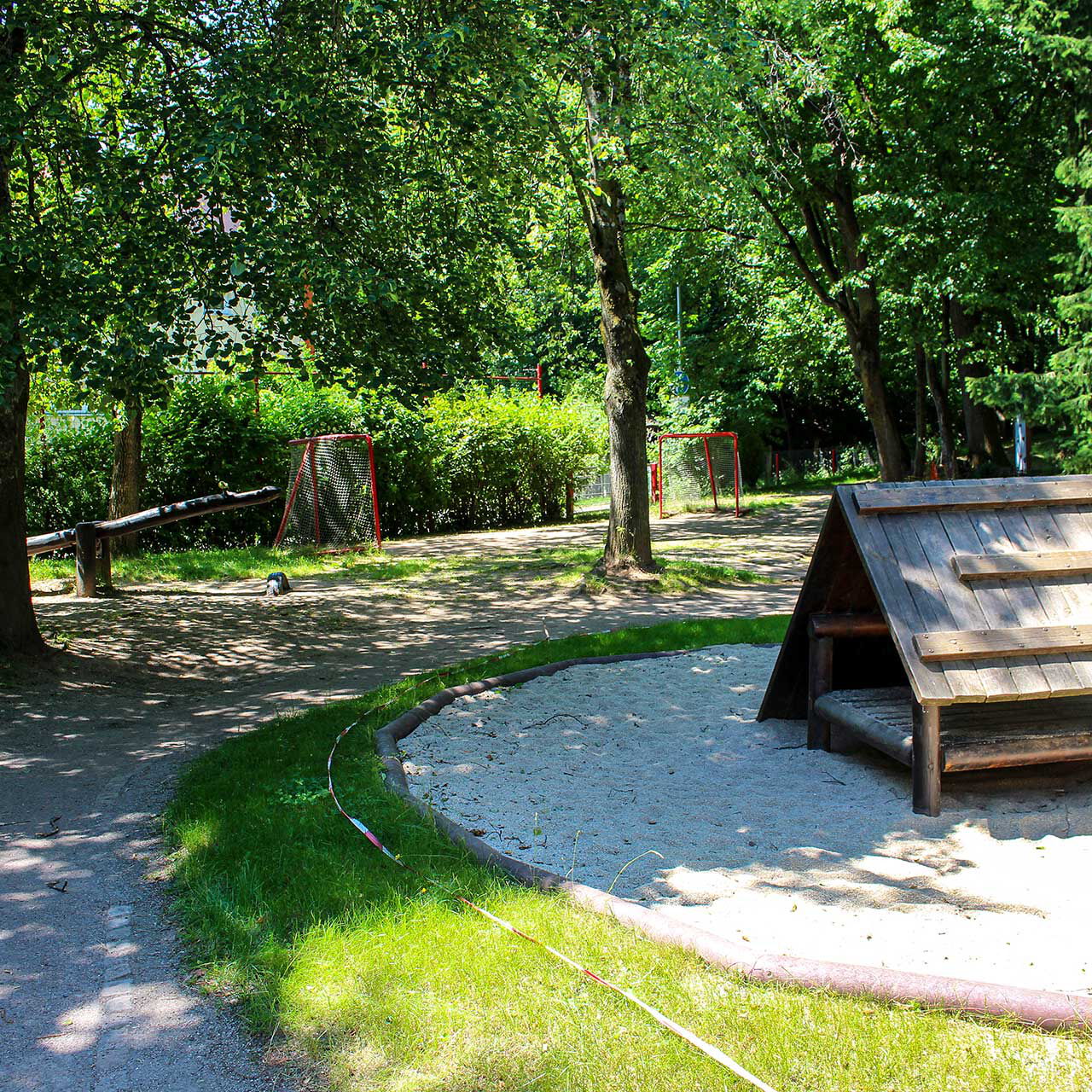Eine Rollerstrecke im Außengelände und in der Mitte steht ein Spielhaus aus Holz. Im Hintergrund stehen zwei kleine Tore am Zaun.