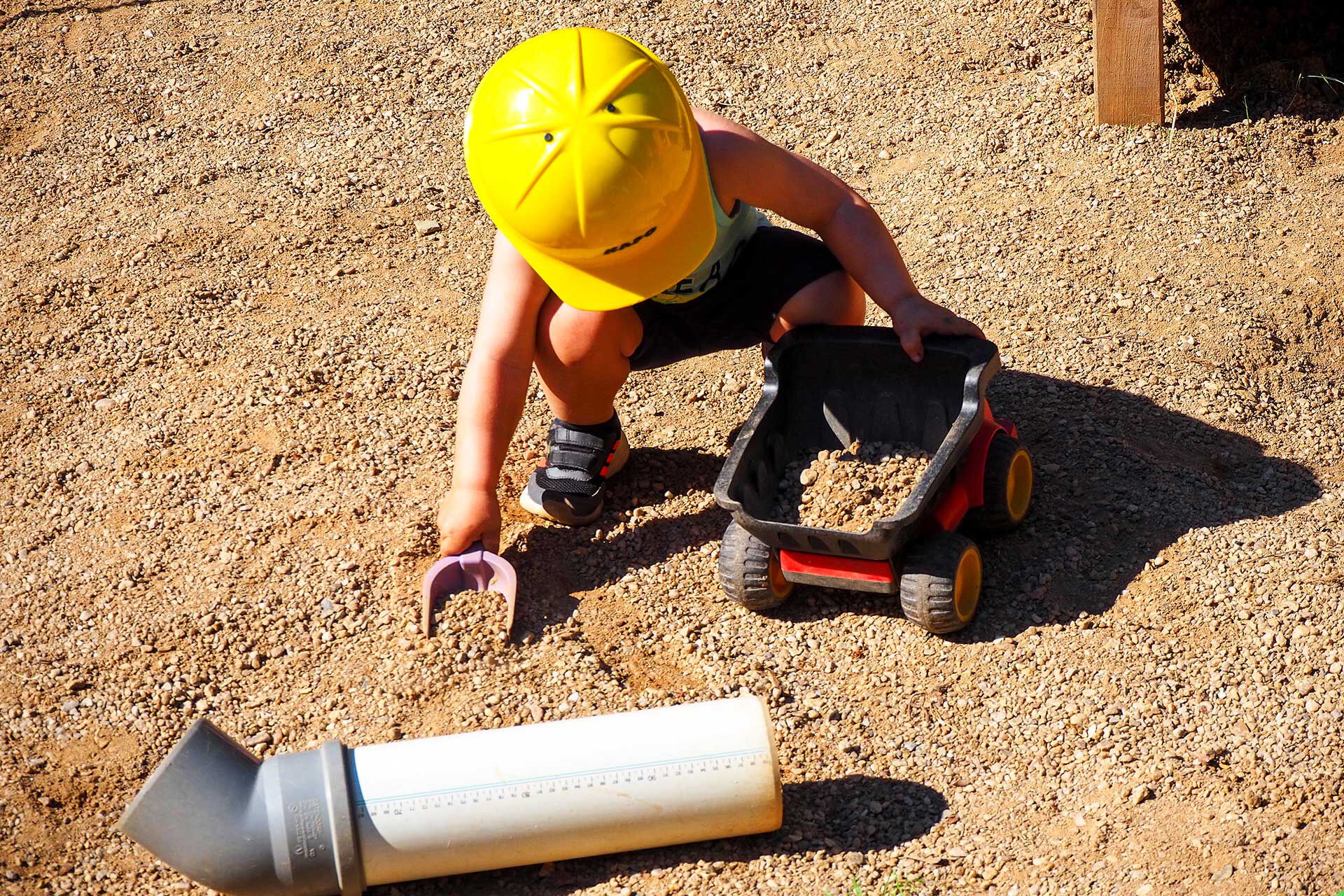 Ein Kleinkind mit einem Bauarbeiterhelm schaufelt Steine und Sand in einen kleinen Plastiklaster.