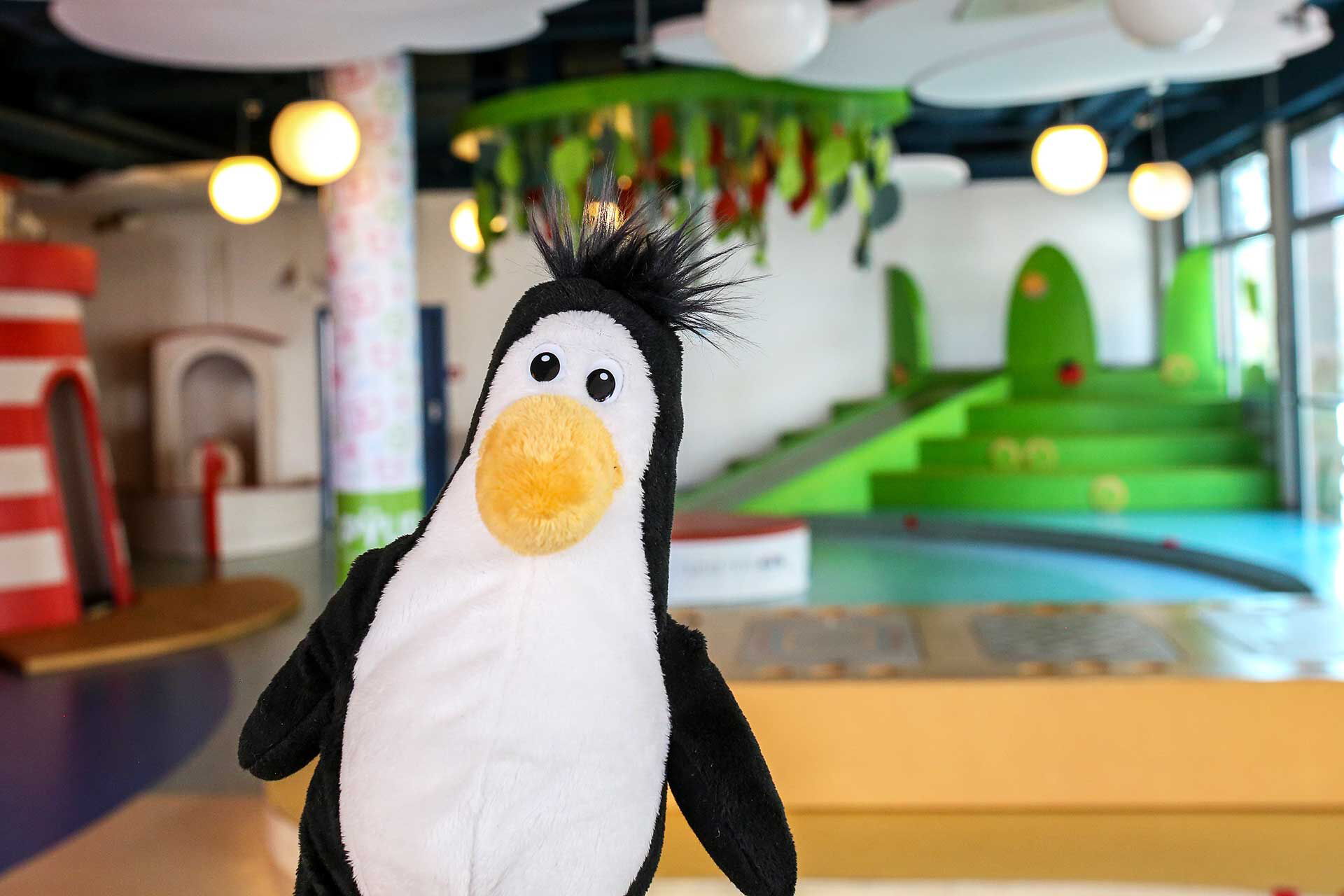 Das Maskottchen vom Pingu Du ist ein Pinguin. Das Plüschtier ist im Vordergrund des Fotos und im Hintergrund ist der Spielbereich zu sehen.