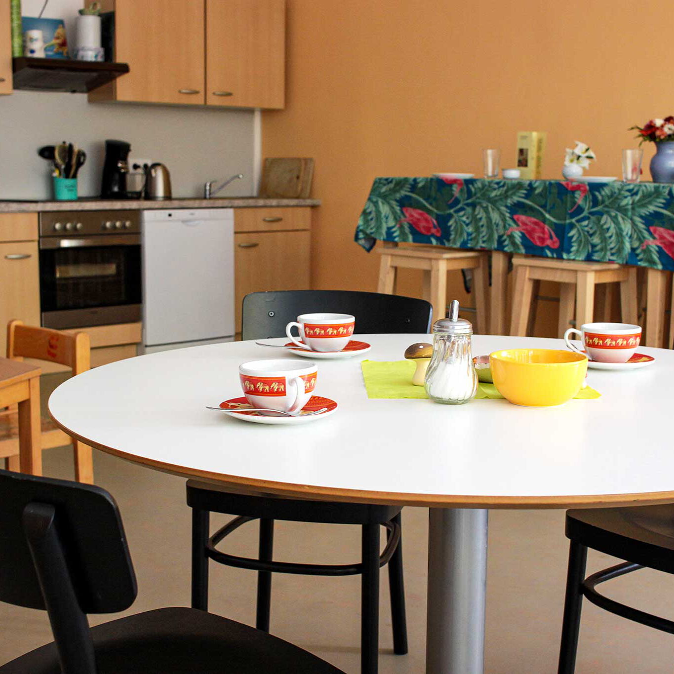 Eine kleine Küchenecke, zwei Tische mit Stühlen und ein Küchentresen mit drei Barhockern.