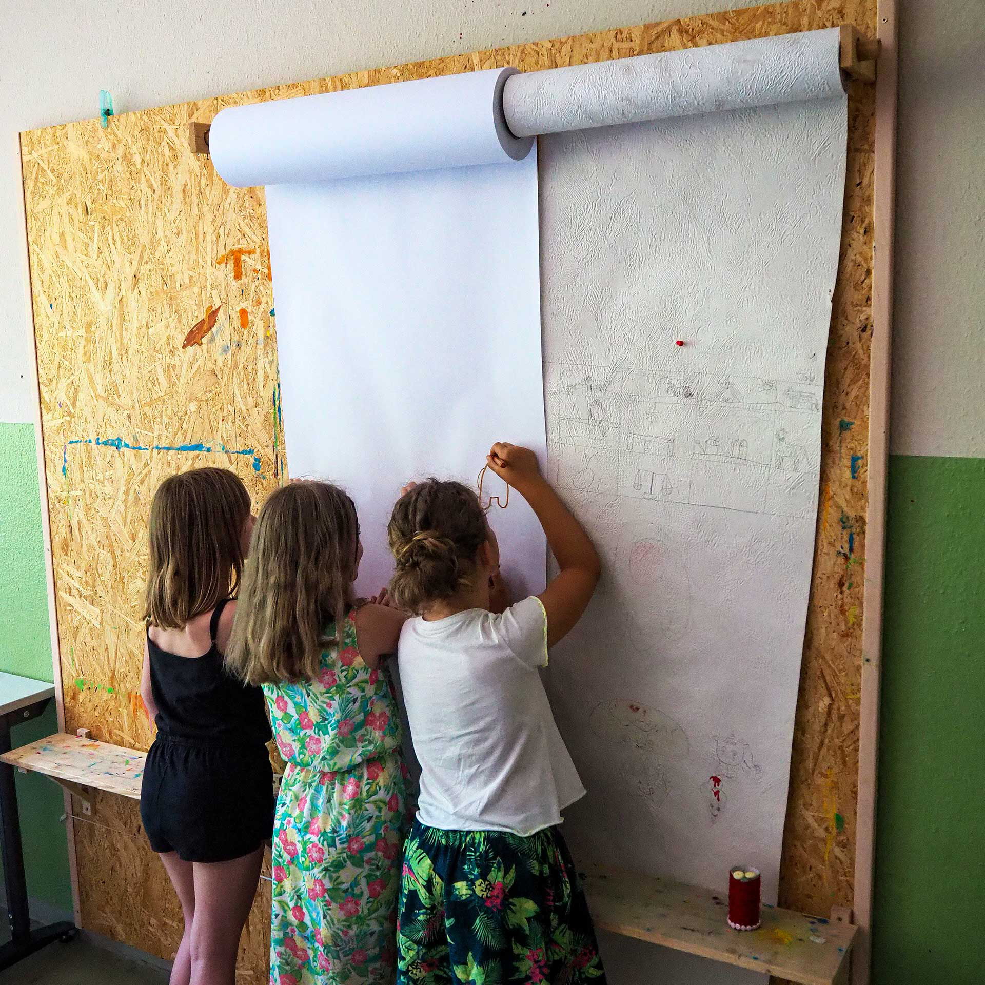 An einer Wand steht eine Spanplatte und an dieser hängen zwei Halterungen für Tapetenrollen. Die Tapeten bemalen drei Kinder.