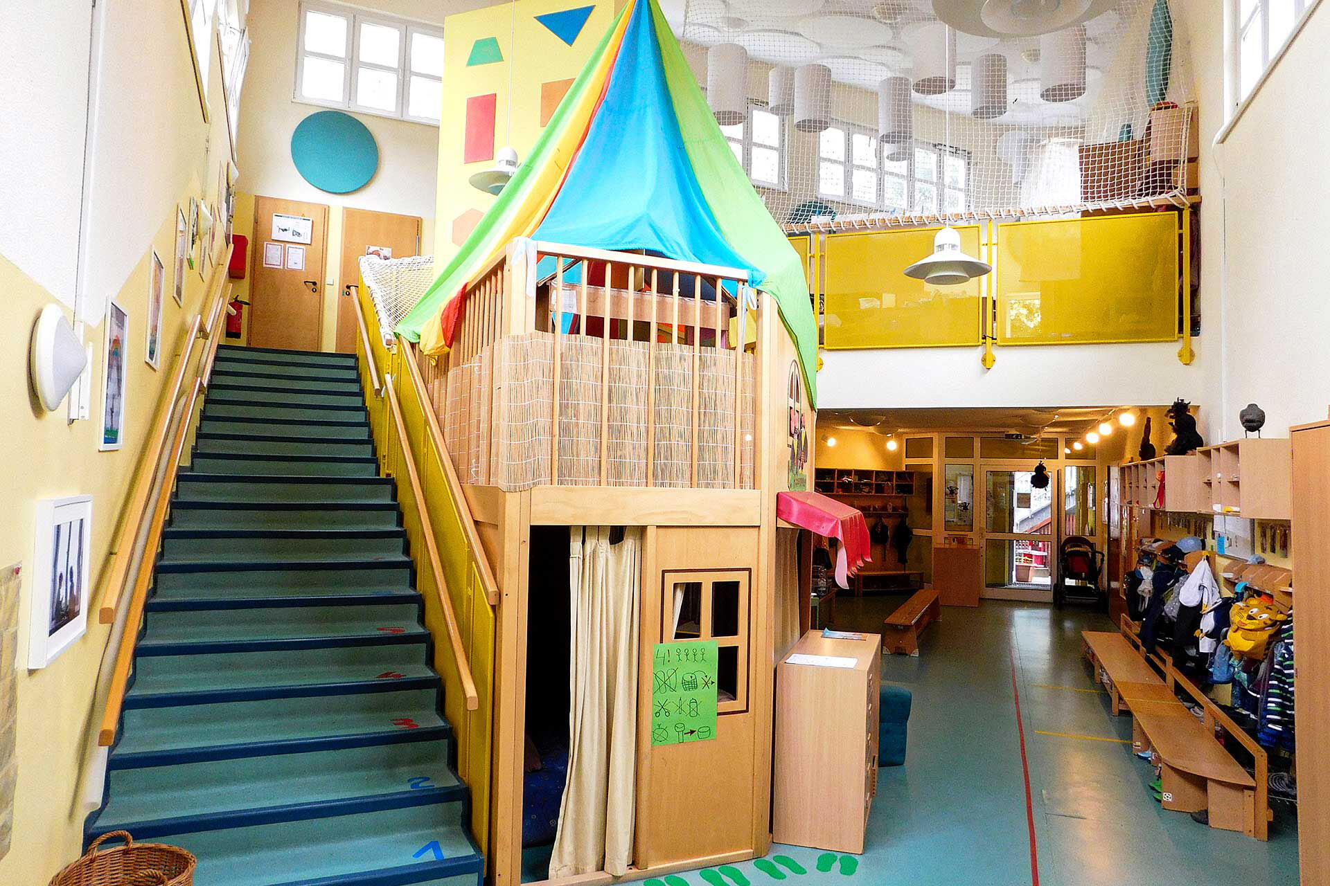 Das offen gestaltete Foyer im Eingangsbereich mit einer offenen Treppe in den oberen Bereich, einem offenen Podest nach unten und einem langen Gang in den unteren Bereich der Kita.