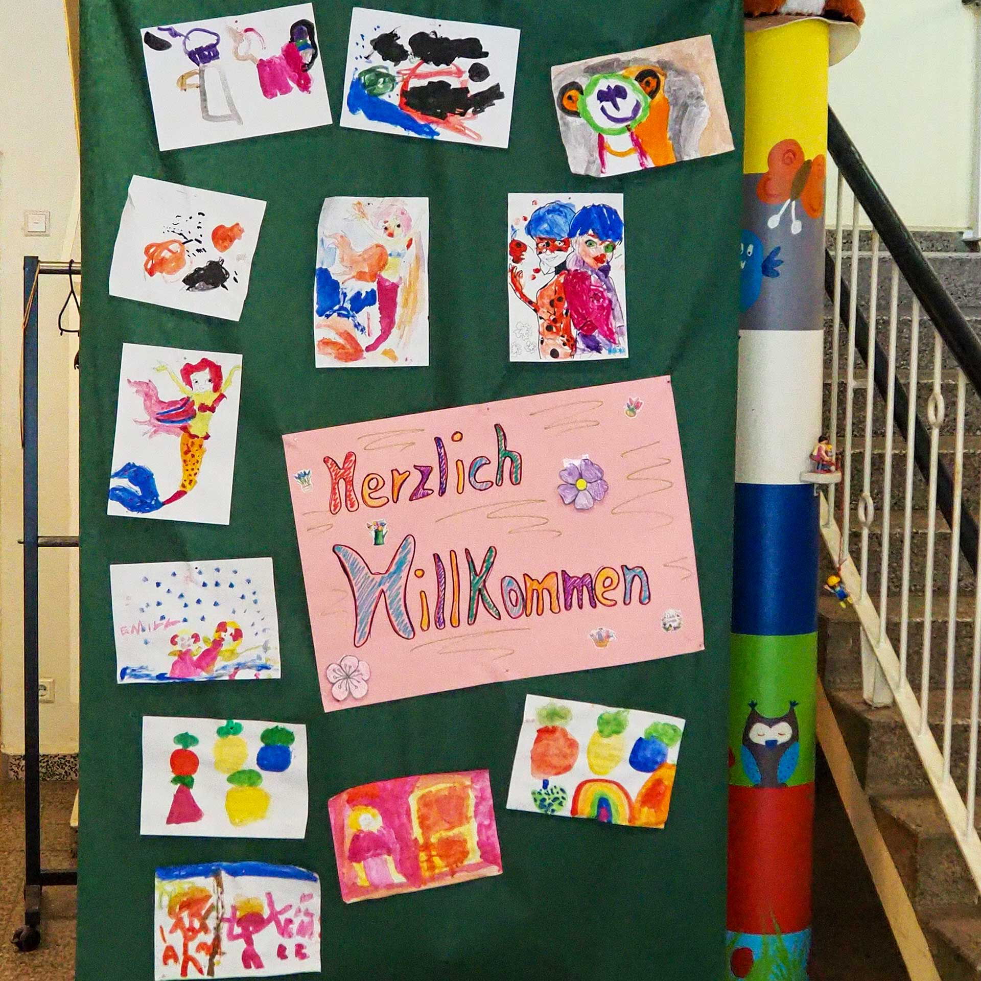 Kinder haben am Haupteingang mehrere Bilder selbst gemalt, auf ein großes grünes Plakat geklebt und in der Mitte steht die Aufschrift Herzlich Willkommen.
