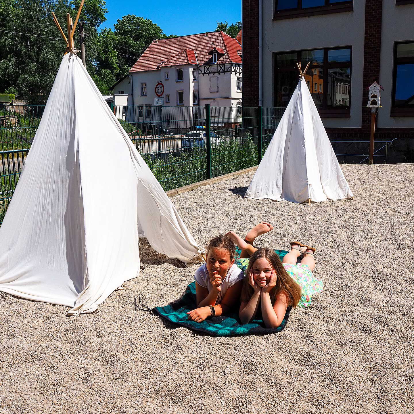 Im Außengeländen stehen zwei Tipis auf dem Fallschutzkies. Im Bildvordergrund liegen zwei Mädchen auf einer Decke vor den Tipis.