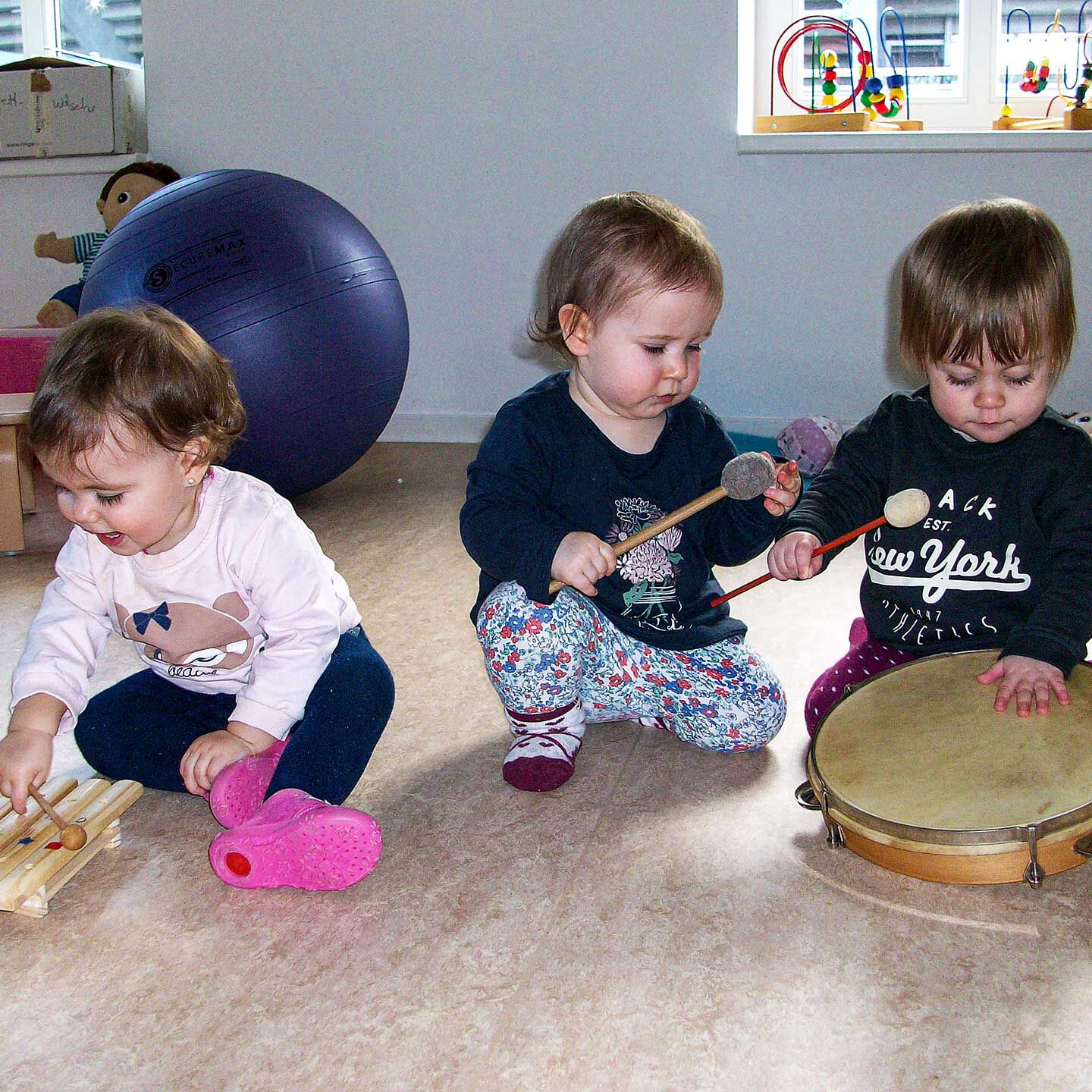 Im Spielzimmer sitzen drei Kleinkinder und musizieren auf einer Trommel und Xylophon.