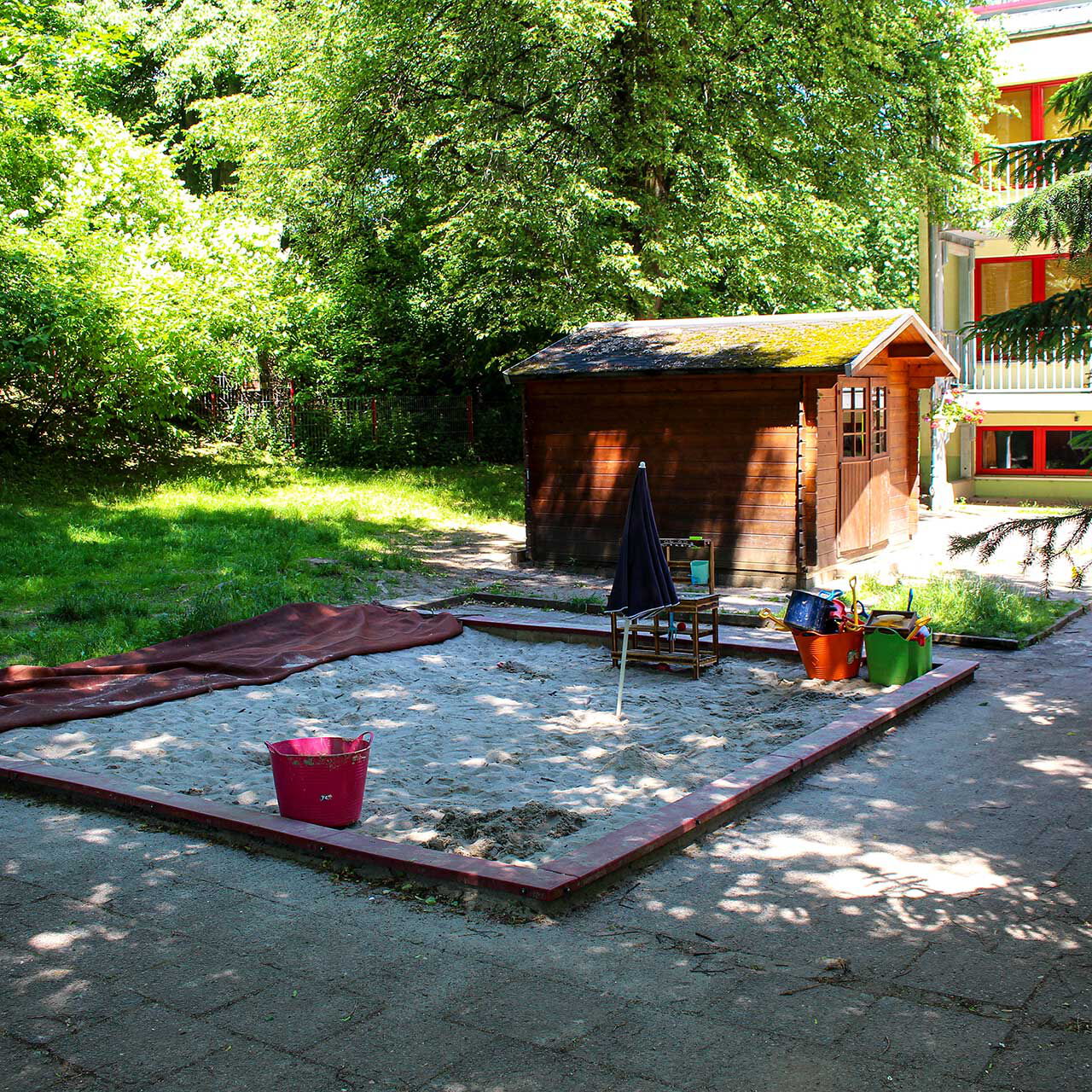 Ein großer Sandkasten im Bildvordergrund und einem Gartenhäuschen im Hintergrund.