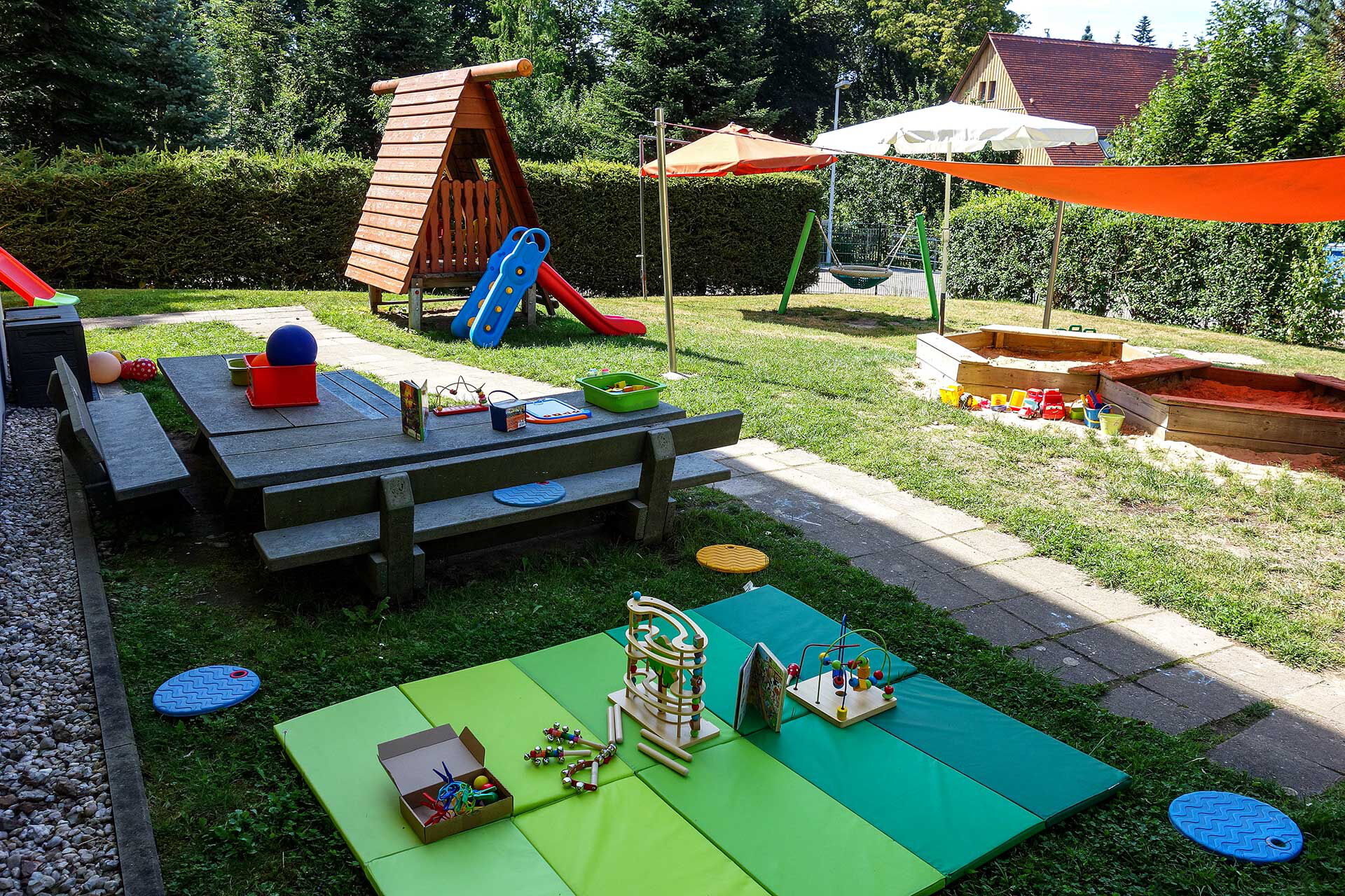 Im Außenbereich ist eine Rutsche, eine Nestschaukel, zwei Sandkästen mit Sonnenschutz und eine Sitzecke.