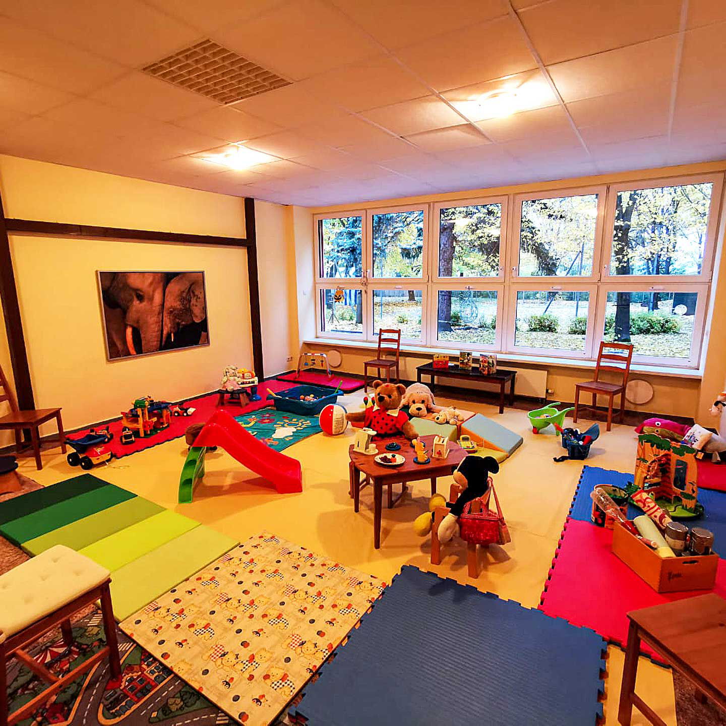 Ein große Spielezimmer mit Rutsche, Spielzeug, Sportmatten, Spielteppiche und Kindertische mit Stühle.