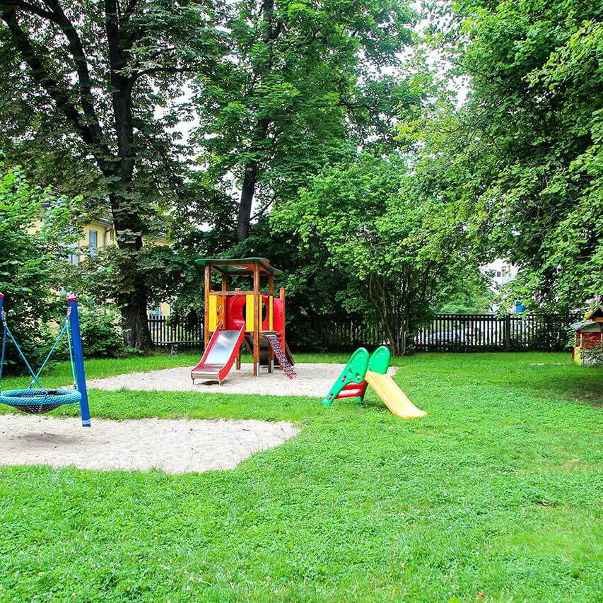 Der Spielplatz im Außengelände mit einer großen Wiese, Nestschaukel, Spielhaus und einem Klettergerüst mit Rutsche.