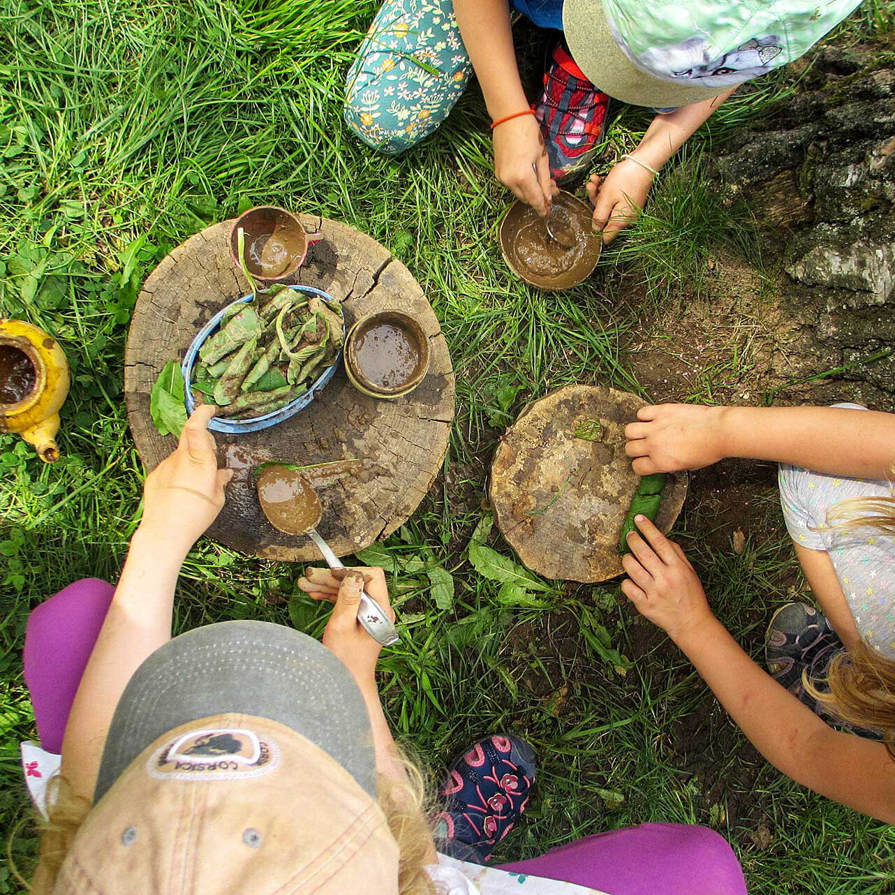 Drei Kleinkinder spielen im Garten mit einer alten Baumscheibe und bereit spielerisch mit Naturmaterialien ein Essen zu.