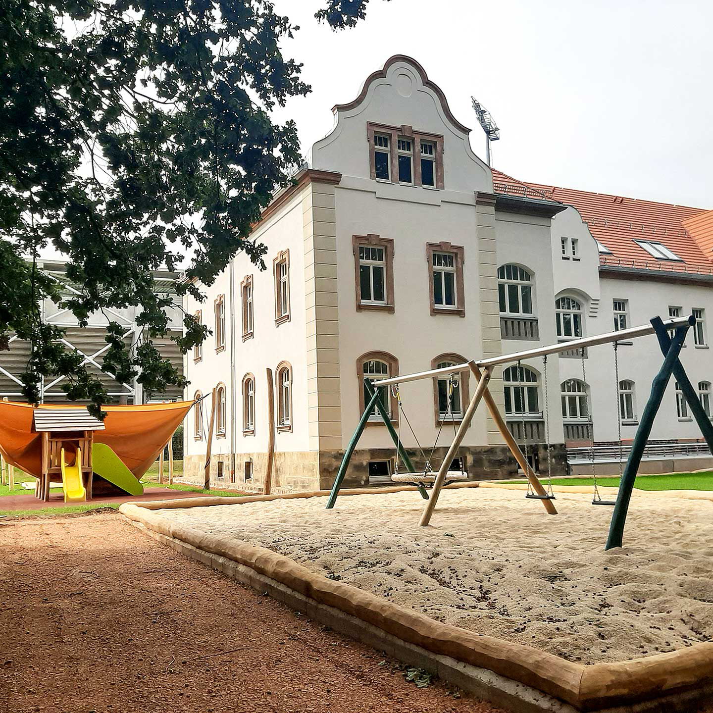 Das Außengelände mit einer Nestschaukel, zwei einzelnen Schaukeln im Vordergrund und das Naturkinderhaus mit einem Spielturm mit Rutsche im Hintergrund.