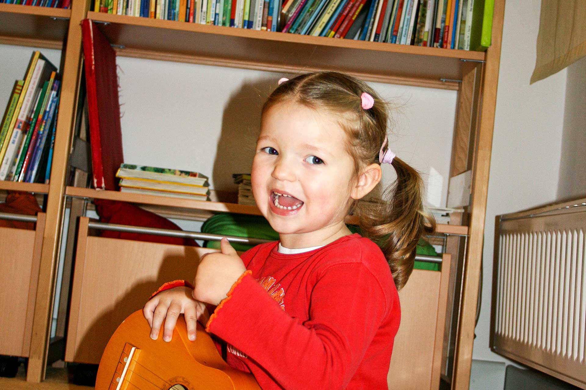 Bücher- und Musikraum Ein Mädchen sitzt auf dem Boden und spielt mit einer kleinen Holzgitarre. Im Hintergrund steht ein Regal mit Büchern.