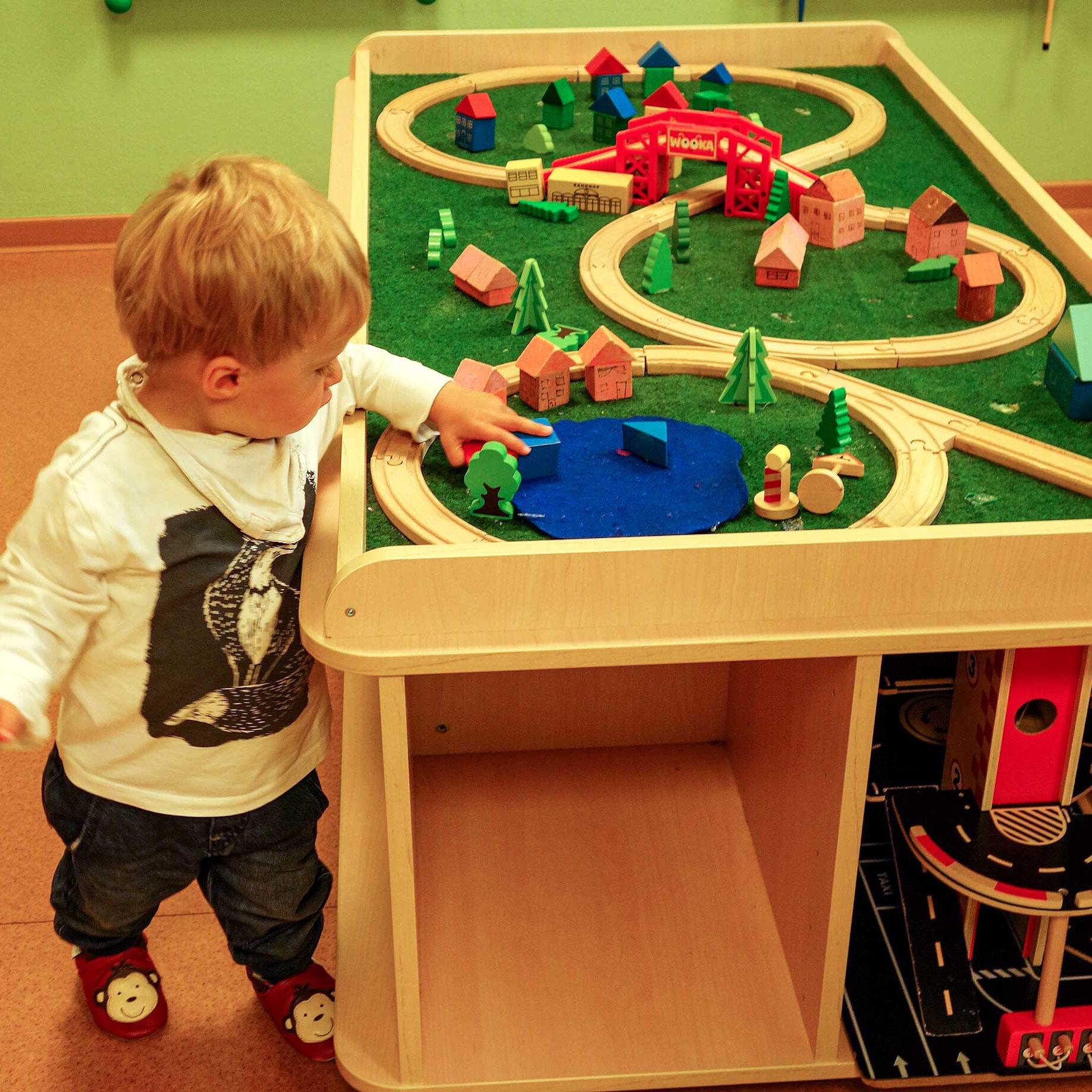 Ein Bautisch auf dem zwei Holzeisenbahnen aufgebaut sind. Am Tisch steht ein Kleinkind und spielt mit den Bausteinen.