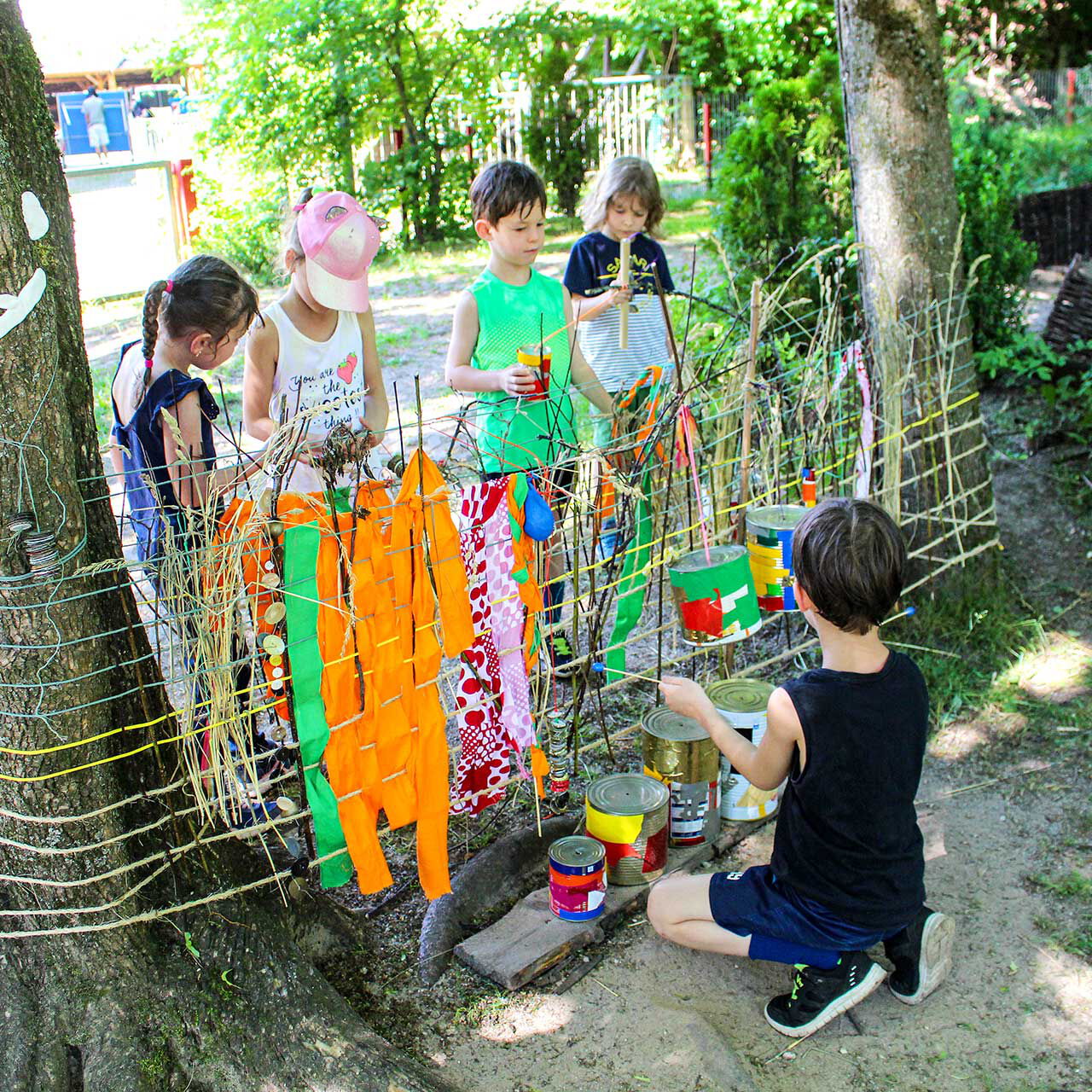 Zwischen zwei Bäumen im Außengelände sind Seile gespannt und Kleidung darüber gehangen. Auf der einen Seite stehen vier Kinder und im Vordergrund kniet ein Kind.