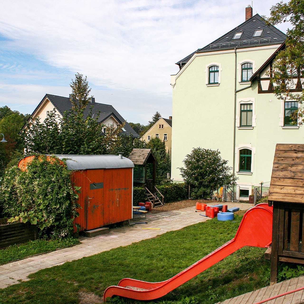 Das Außengelände der Kita mit Rutsche, Klettergerüst, Spielhaus, Bauwagen und Hochbeeten.