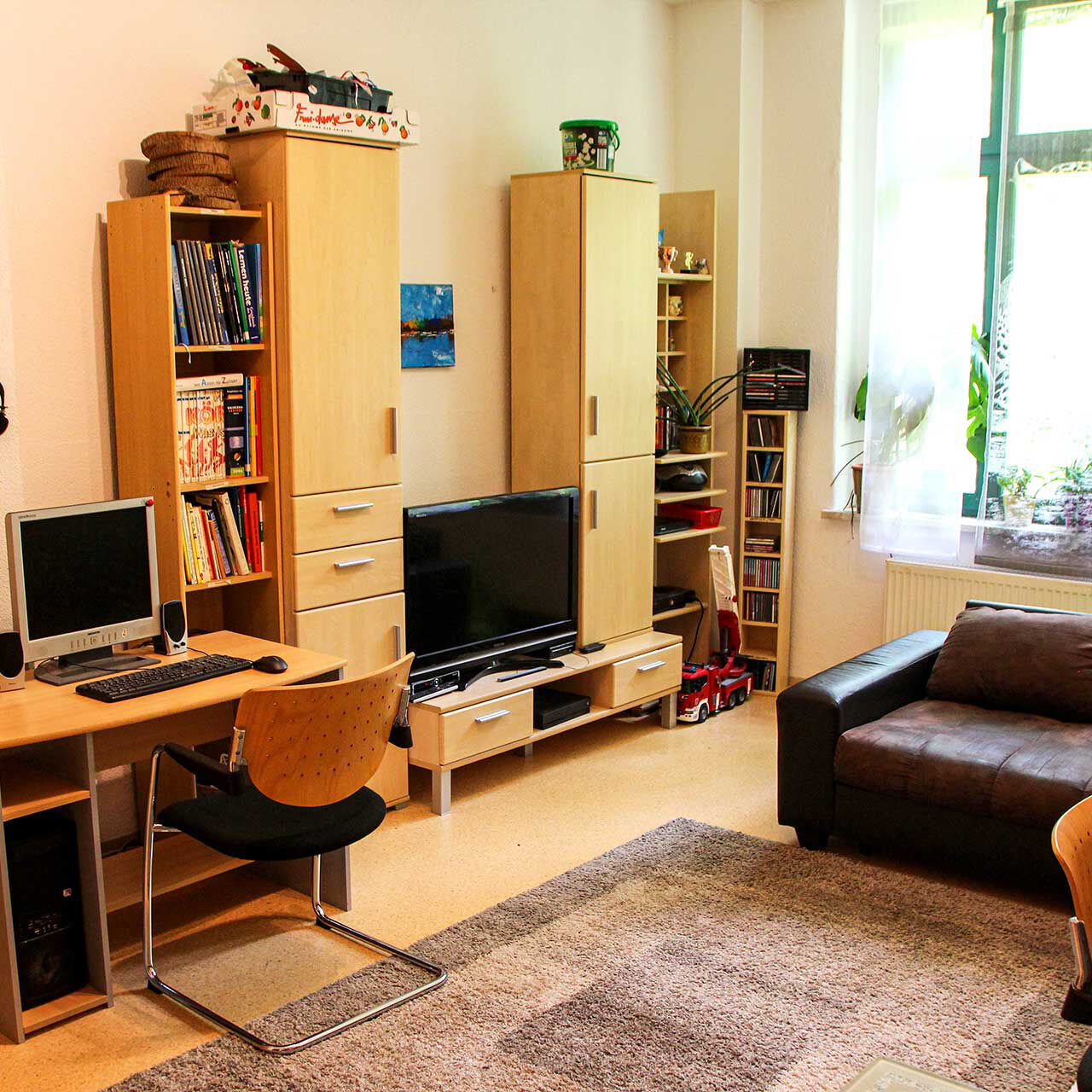 Das Gruppenzimmer mit einem Ecksofa, Schreibtisch mit PC, einer Anbauwand und einem TV.