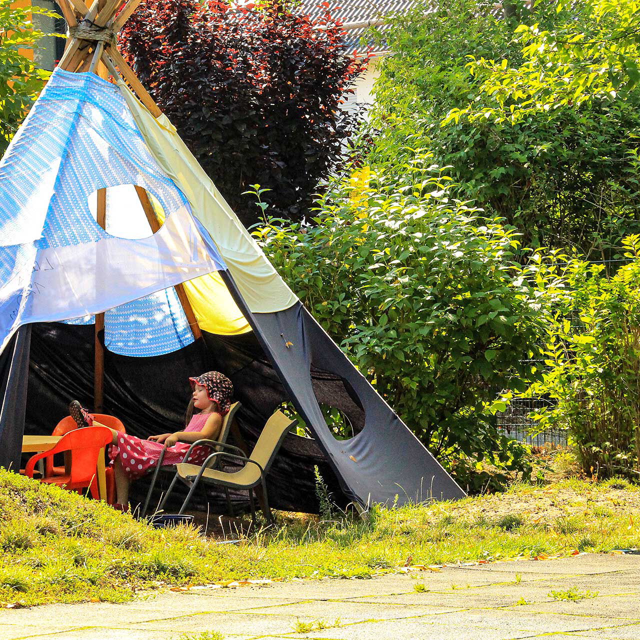 Ein Tipi steht auf der Wiese im Außengelände. Zwei Kleinkinder spielen im und am Zelt.