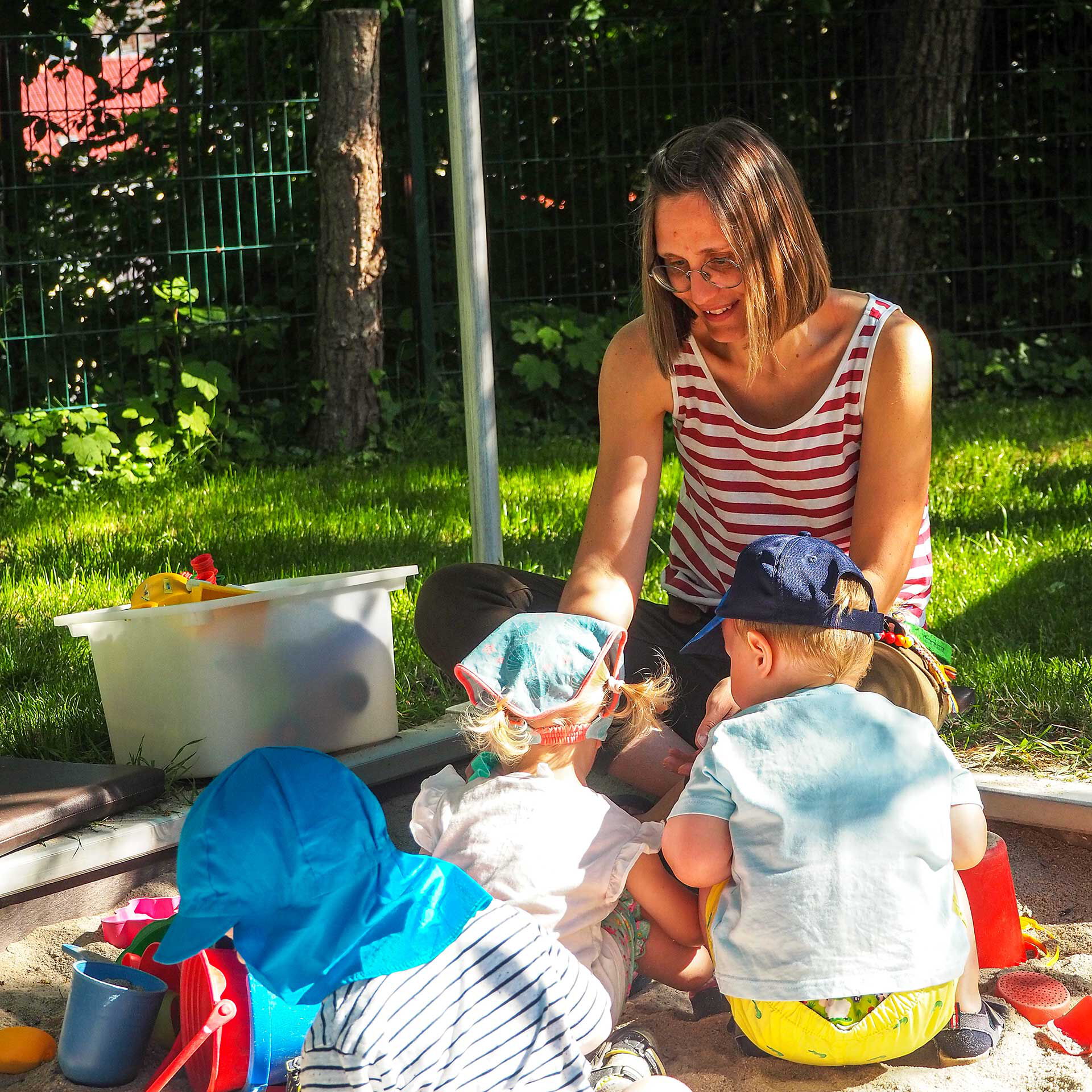 Drei Kinder sitzen mit ihrer Erzieherin im Sandkasten und spielen mit dem Sandspielzeug.