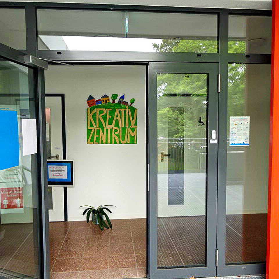 Der Eingangsbereich des Kreativzentrums bestehend aus einer vollverglasten grauen Eingangstür, einem Schriftzug Kreativzentrum auf der rechten Seite und einem selbst gemalten Bild des Kreativzentrums im Bildmittelpunkt.
