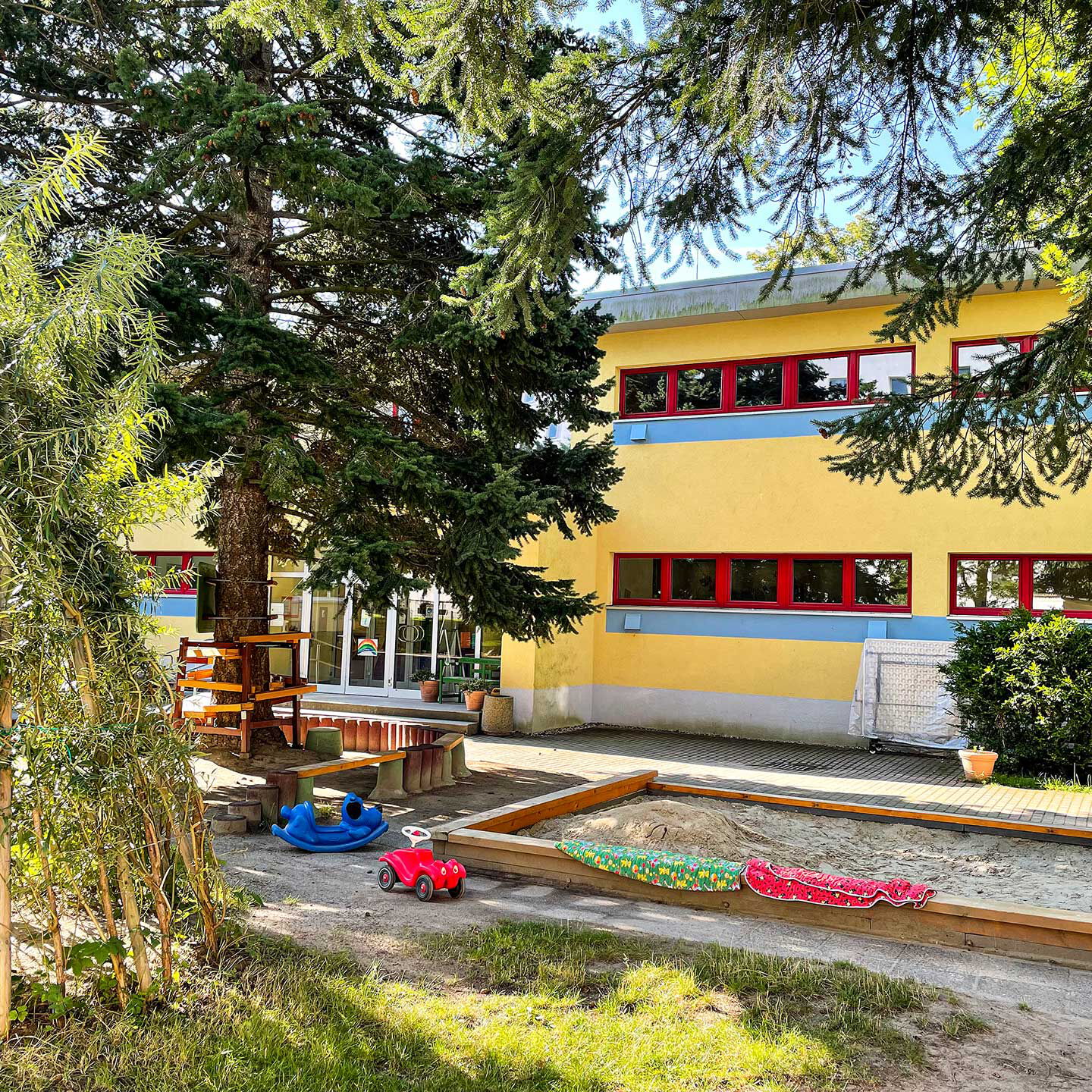 Das Außengelände im Eingangsbereich mit einem Sandkasten, einem Tipi aus Weide und einem Sitzbereich unter einem Baum.