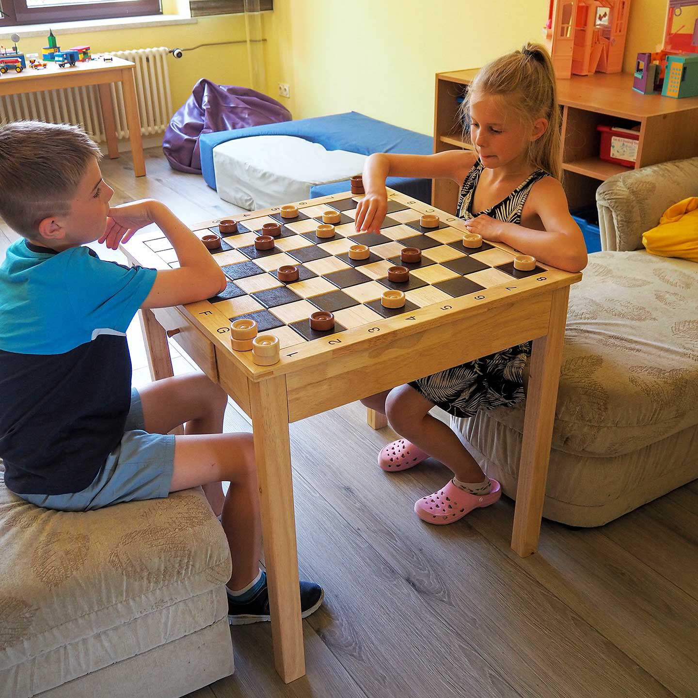 Ein Schachbretttisch im Spielzimmer an denen zwei Kinder Dame spielen.