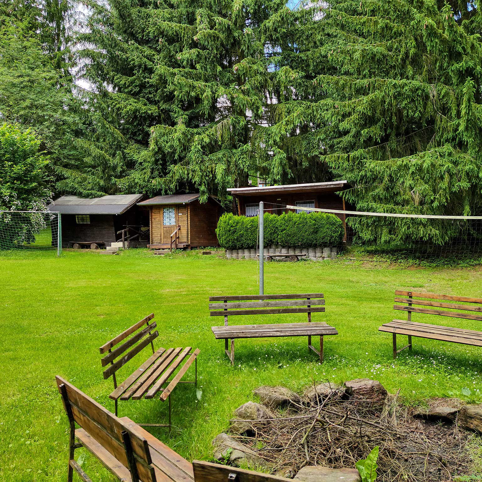 Im Vordergrund der Lagerfeuerplatz mit 5 Holzbänken, im Hintergrund der Volleyballplatz und 3 Holzblockhütten.