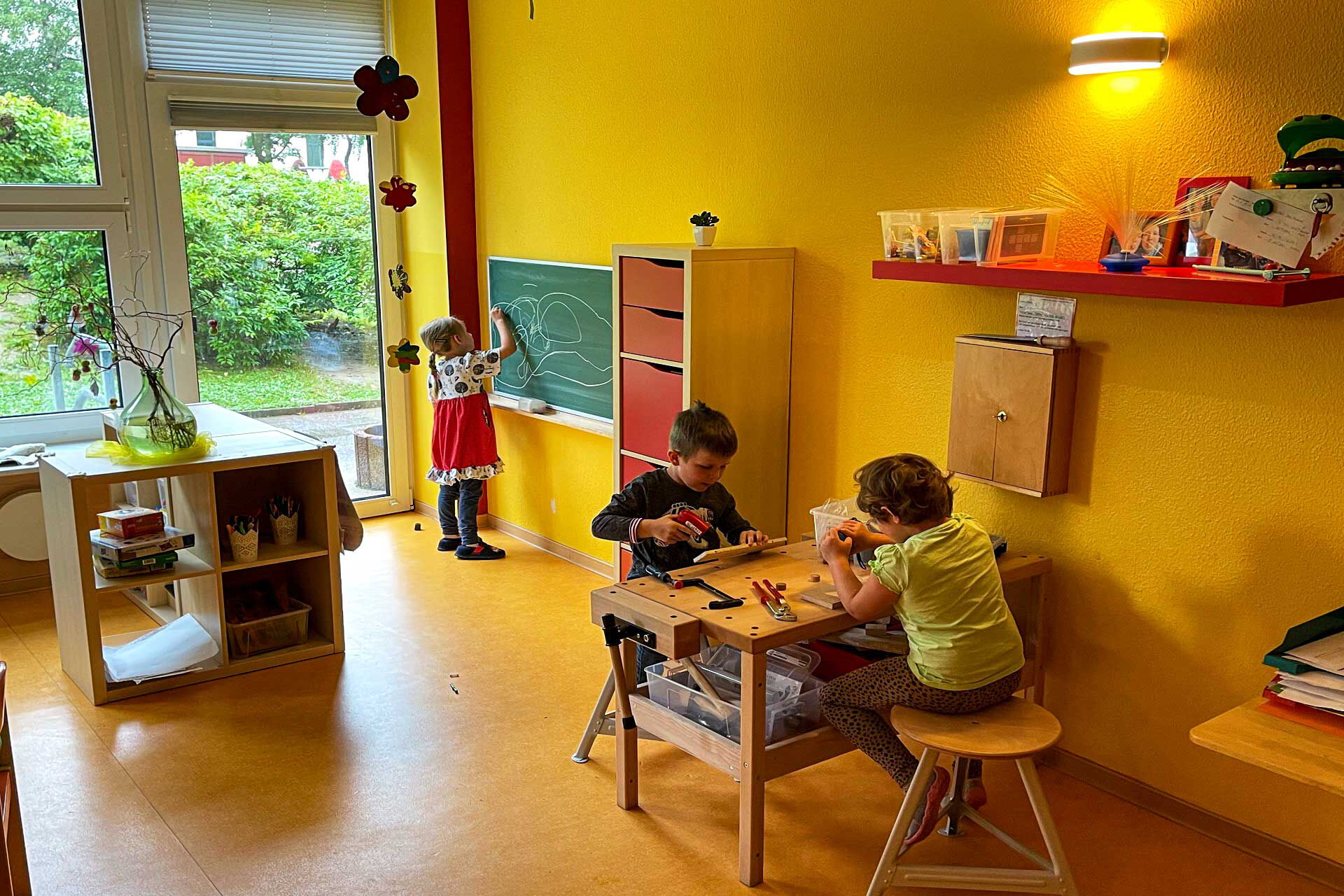 Eine Werkbank steht an der Wand in einem Zimmer und zwei Kinder sitzen auf Stühlen an der Werkbank. Ein Mädchen malt mit Kreide an eine Wandtafel.