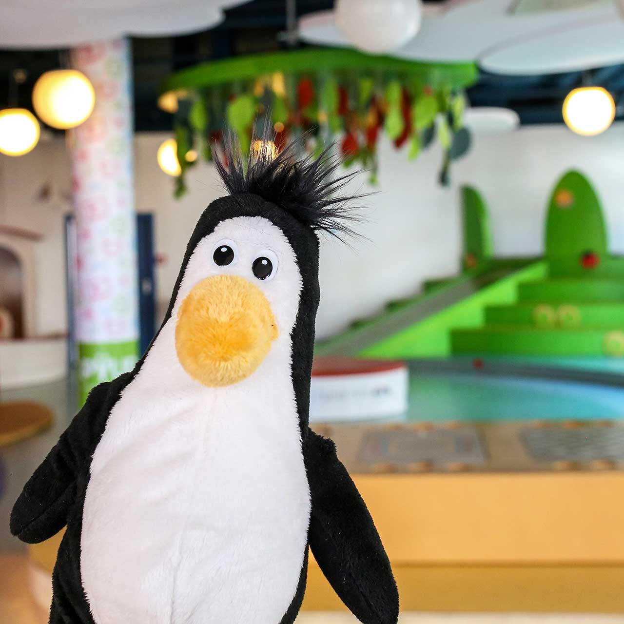 Das Maskottchen vom Pingu Du ist ein Pinguin. Das Plüschtier ist im Vordergrund des Fotos und im Hintergrund ist der Spielbereich zu sehen.