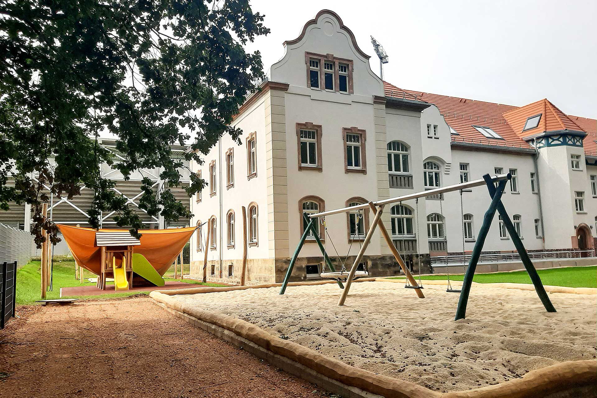 Das Außengelände mit einer Nestschaukel, zwei einzelnen Schaukeln im Vordergrund und das Naturkinderhaus mit einem Spielturm mit Rutsche im Hintergrund.