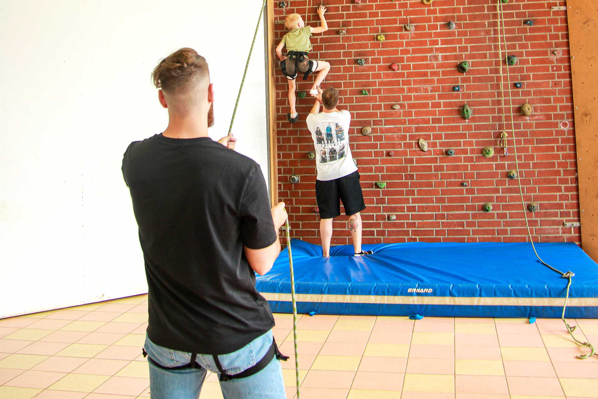 An der Kletterwand im Jugendhaus Compact klettert ein Kind hoch und wird von zwei Pädagogen gestützt bzw. gesichert.