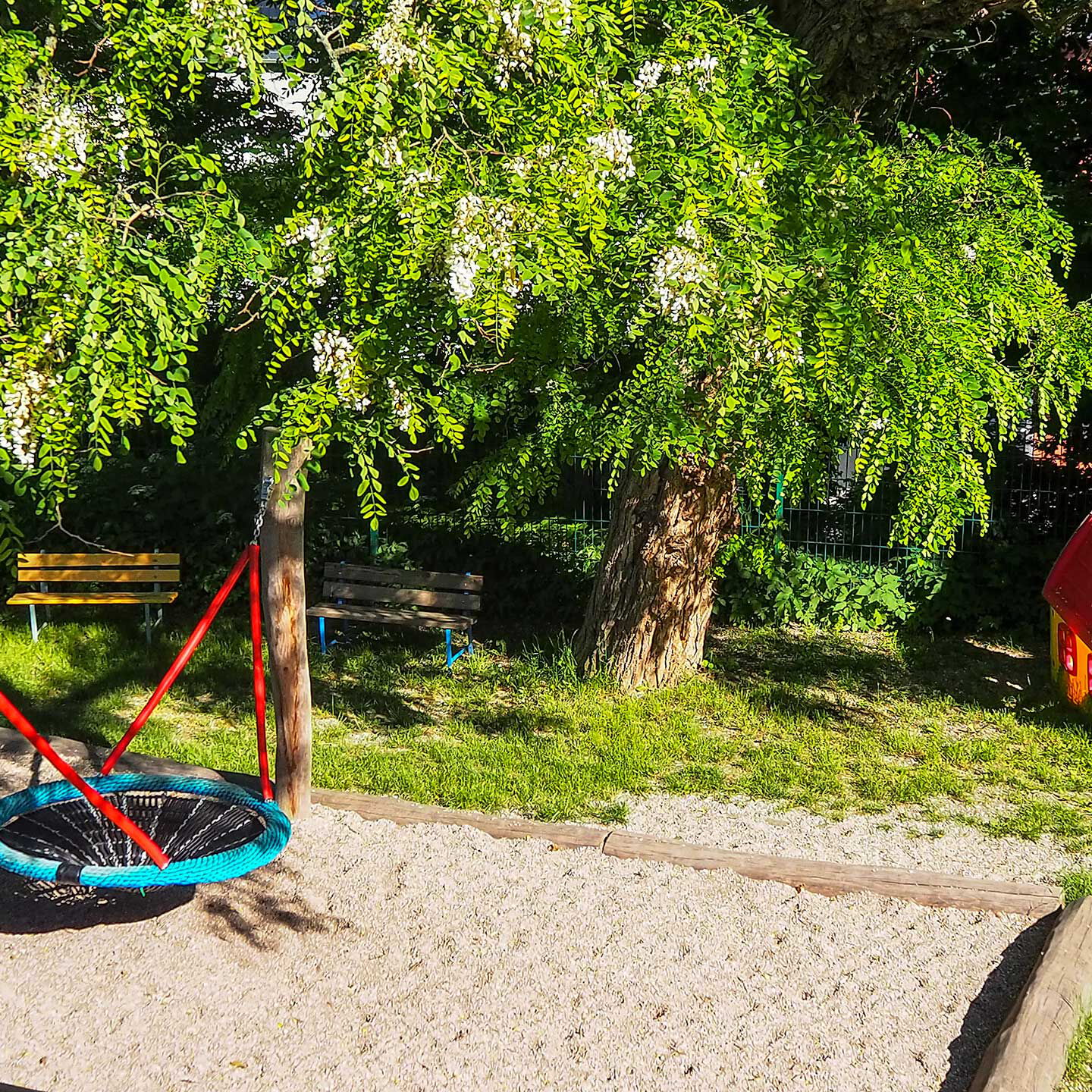 Eine Nestschaukel im Außenbereich unter einem Baum. Am rechten Bildrand steht ein kleines Spielhaus aus Plastik.