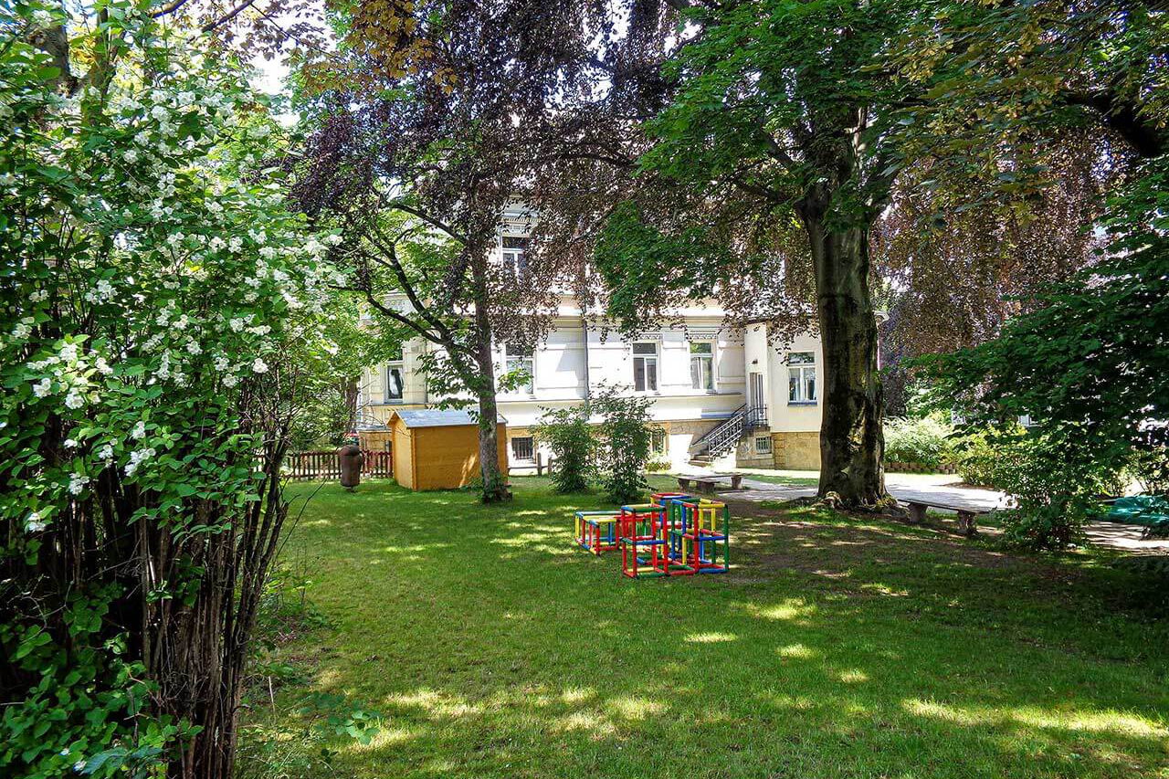 Der Gartenbereich mit einer großen Wiese, vielen Bäumen, einer Laube und einem transportablen Klettergerüst.