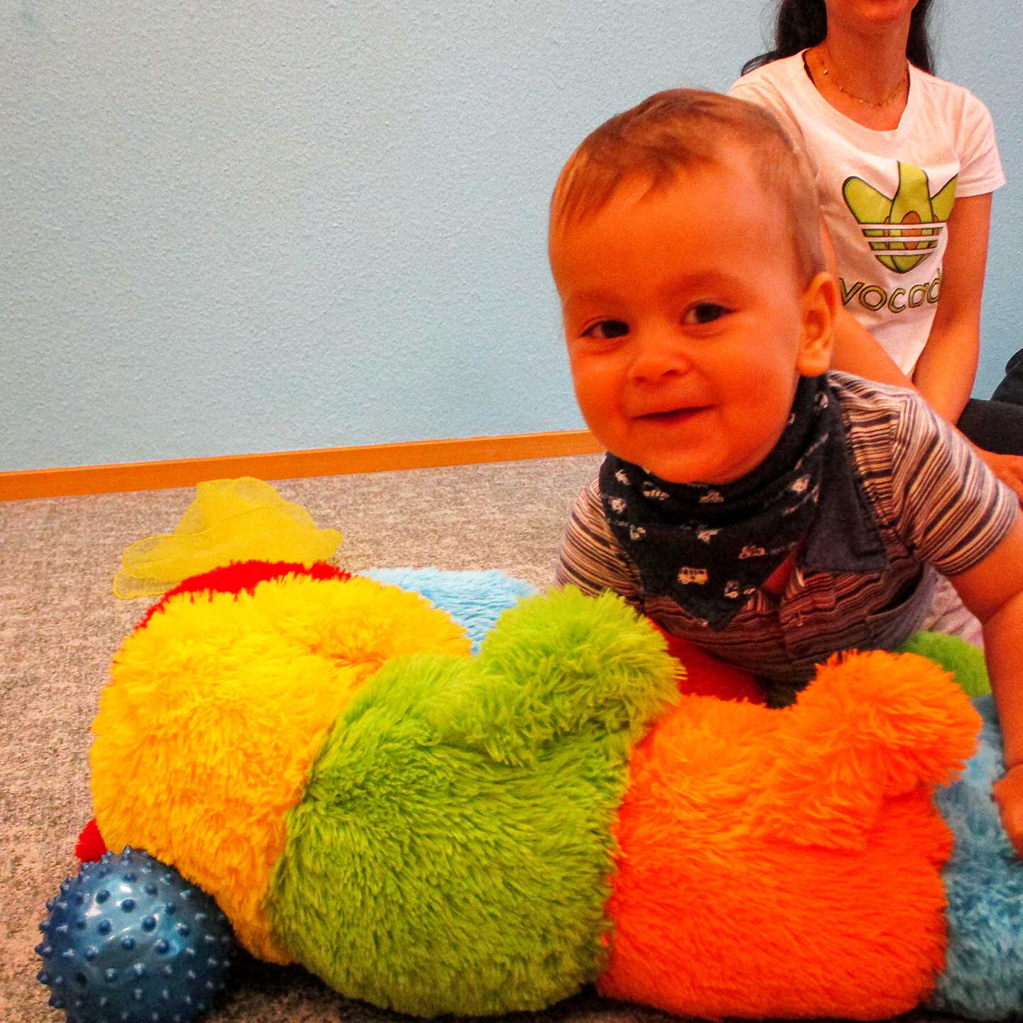 Ein Kleinkind spielt mit einem Kuscheltier und im Hintergrund sitzt seine Mama.