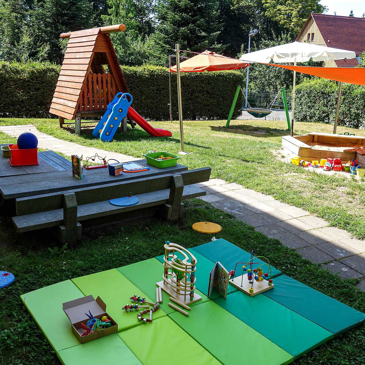 Im Außenbereich ist eine Rutsche, eine Nestschaukel, zwei Sandkästen mit Sonnenschutz und eine Sitzecke.