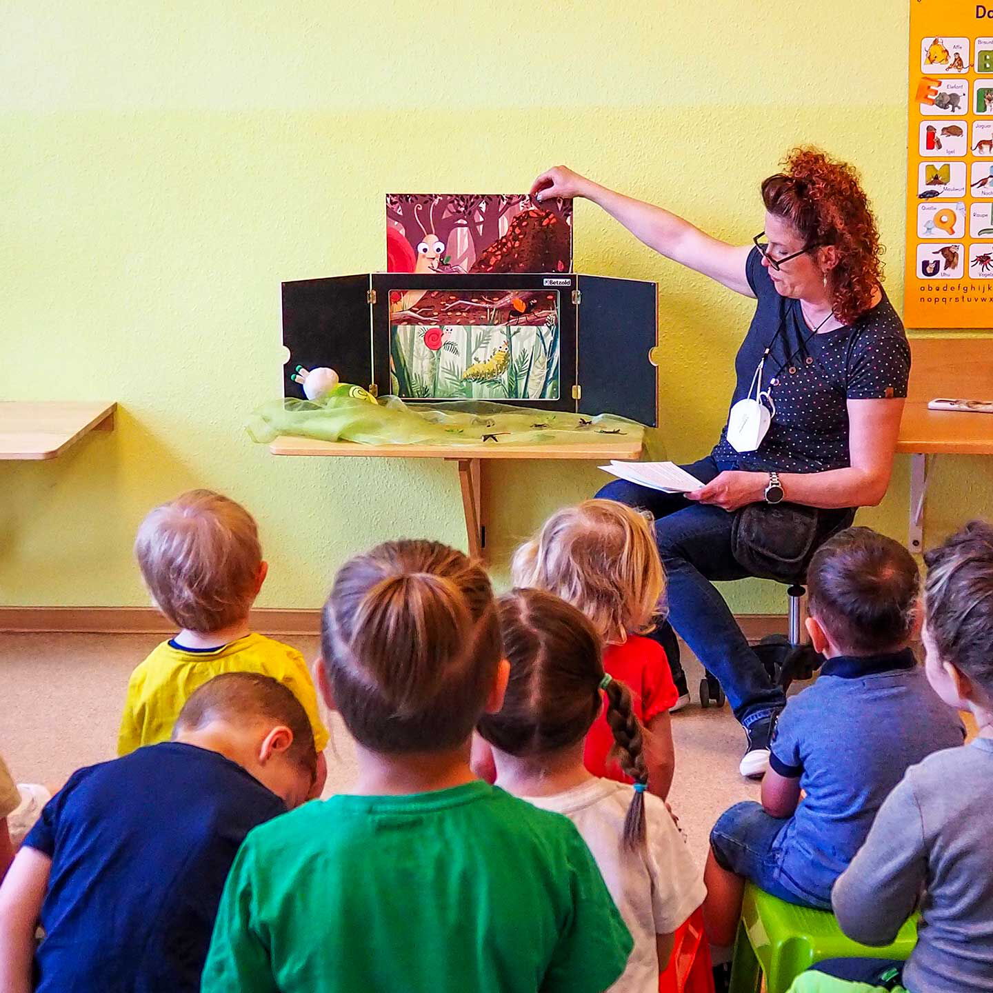 Eine Gruppe von Kindern sitzt vor einer kleinen Leinwand. Eine Mitarbeiterin liest eine Geschichte und schiebt unterschiedliche Bilder in die Leinwand.