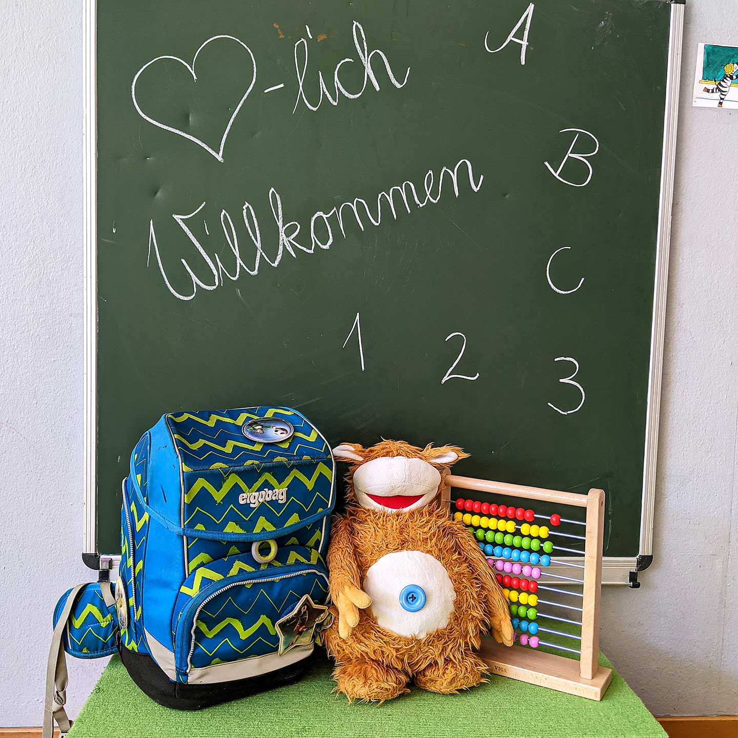 Auf einem Tisch stehen ein Ranzen, Rechenschieber und Kuscheltier. Dahinter steht auf einer Wandtafel Herzlich Willkommen.