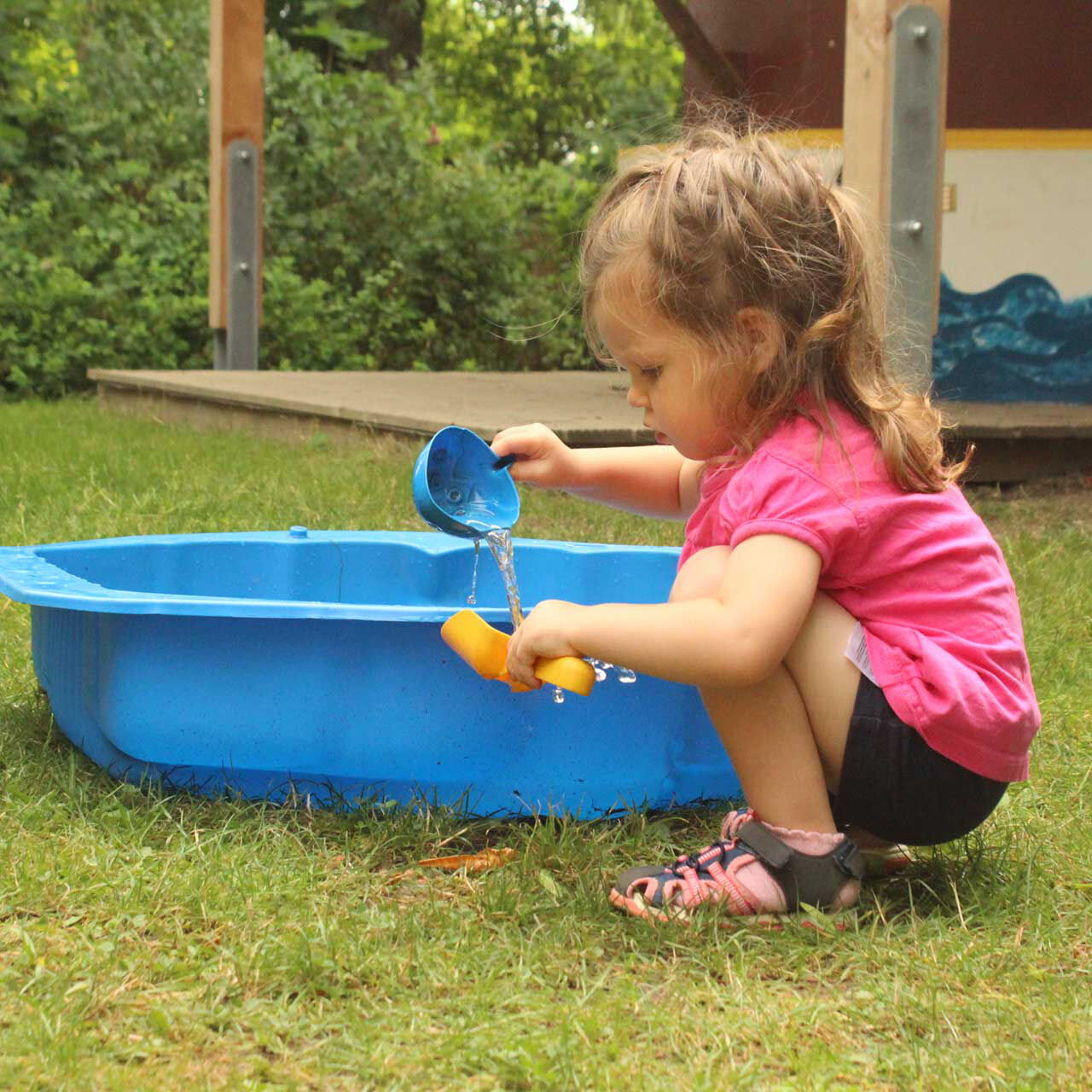 Ein Kleinkind spielt mit einer Schaufel im Garten und nutzt dazu das Wasser aus einer blauen Plastikwanne.