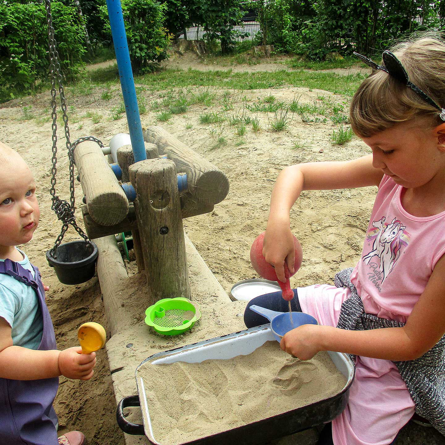 Im Sandkasten sitzen zwei Kleinkinder und spielen mit Schaufeln an einem Holzbagger.