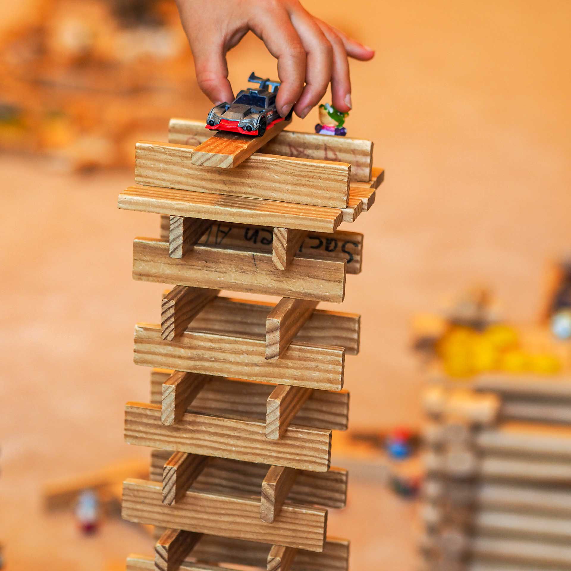 Ein selbst gebauter Turm aus flachen Holzbausteinen. Auf dem Dach wird von einem Kind ein Spielzeugauto abgestellt.
