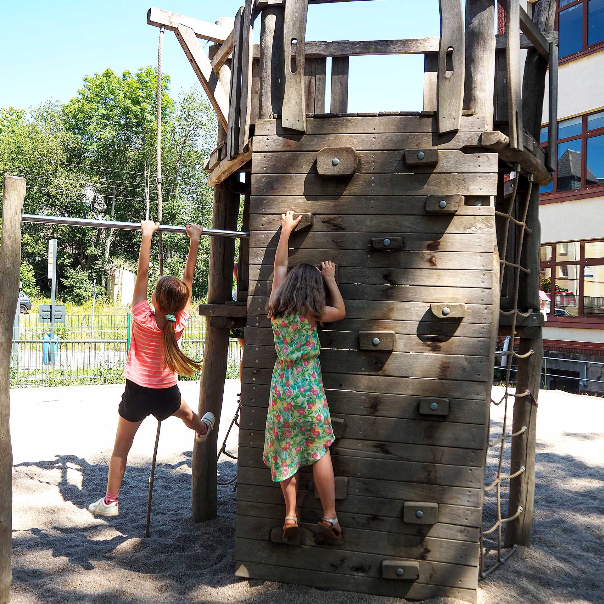 Ein Kletterturm aus Holz mit vielen verschiedenen Klettermöglichkeiten. Zwei Mädchen klettern gerade den Turm hinauf.