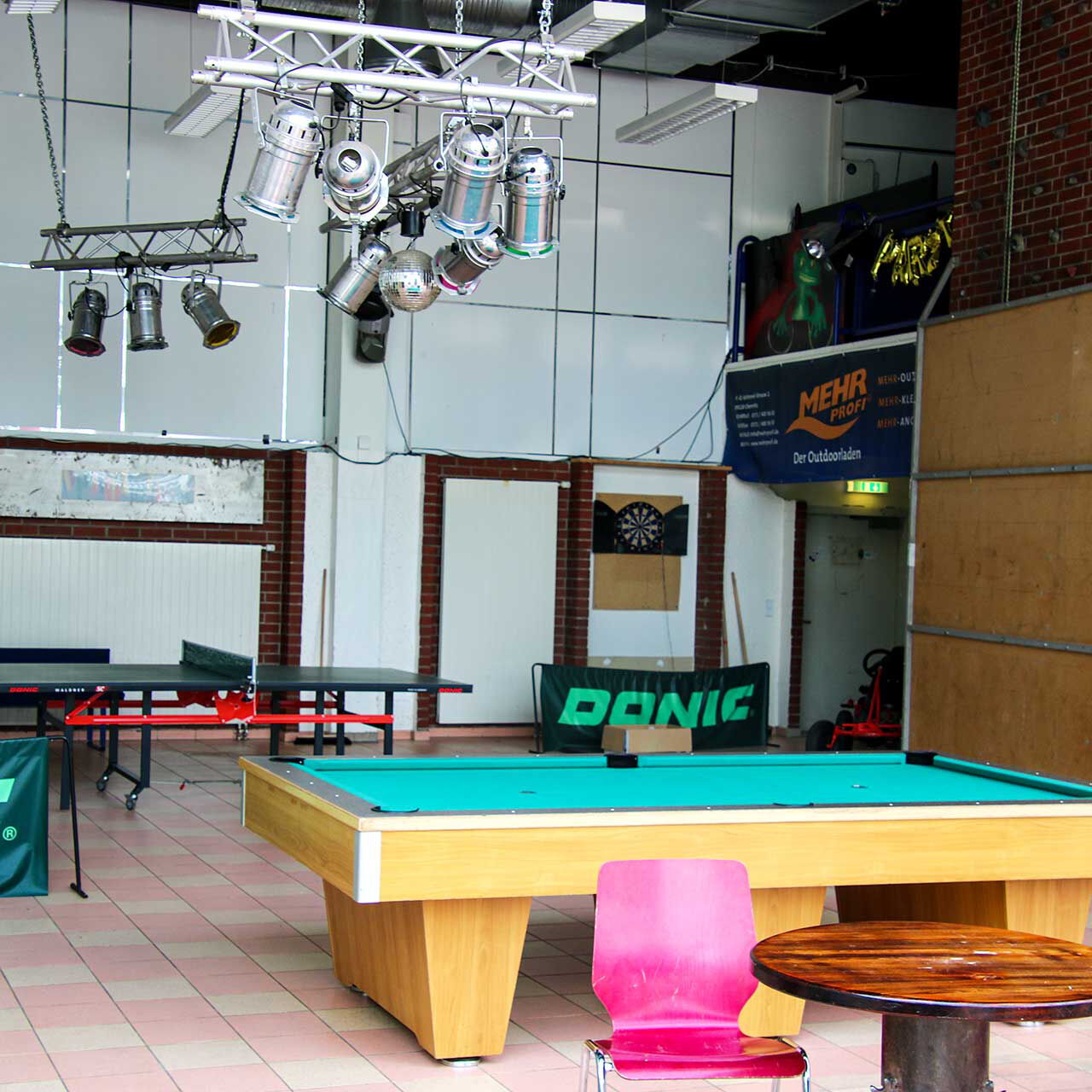 Die Spielhalle mit einer Tischtennisplatte, dem Billardtisch, DJ-Bereich und einem Dartspiel.