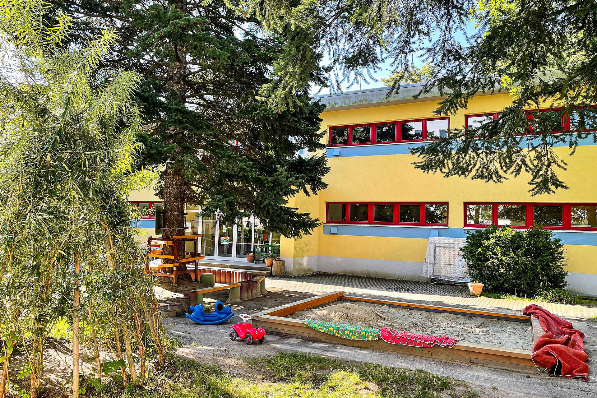 Der Außenbereich mit einem Sandkasten, Sitzbereich, Spielzeug und Baumhöhle.