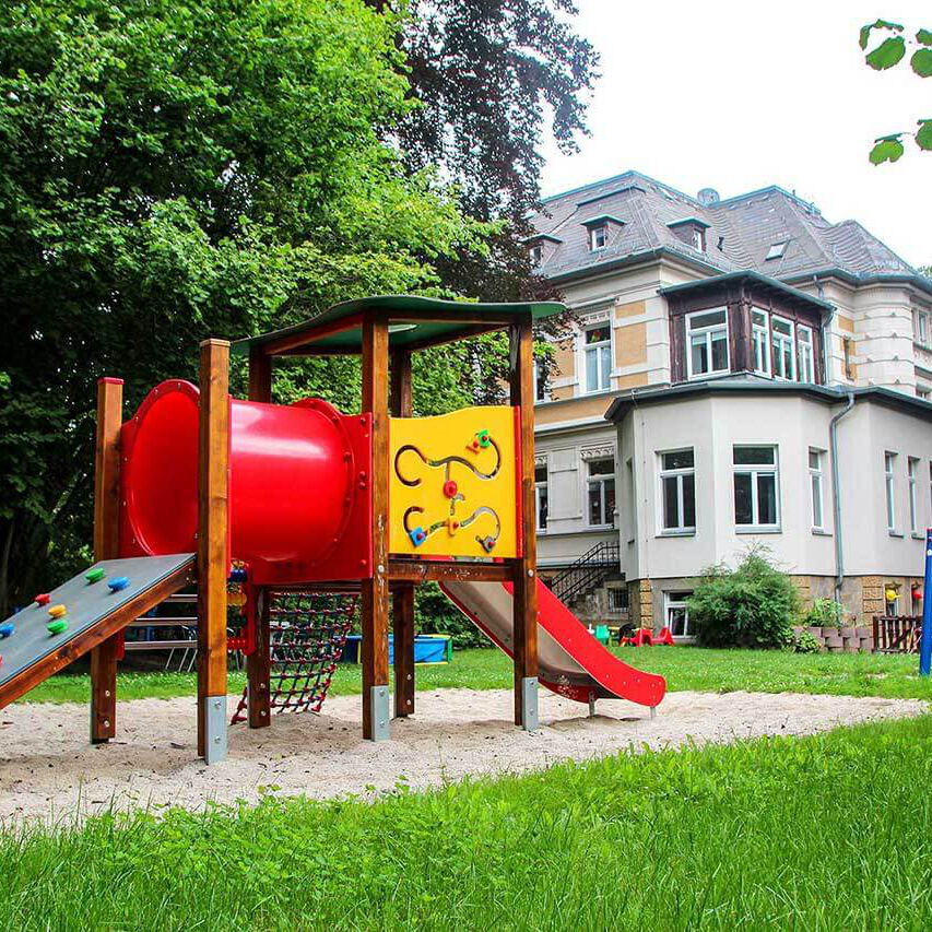 Der Spielplatz im Außengelände mit einem Klettergerüst mit Rutsche und einer Nestschaukel. Im Hintergrund ist die Villa.