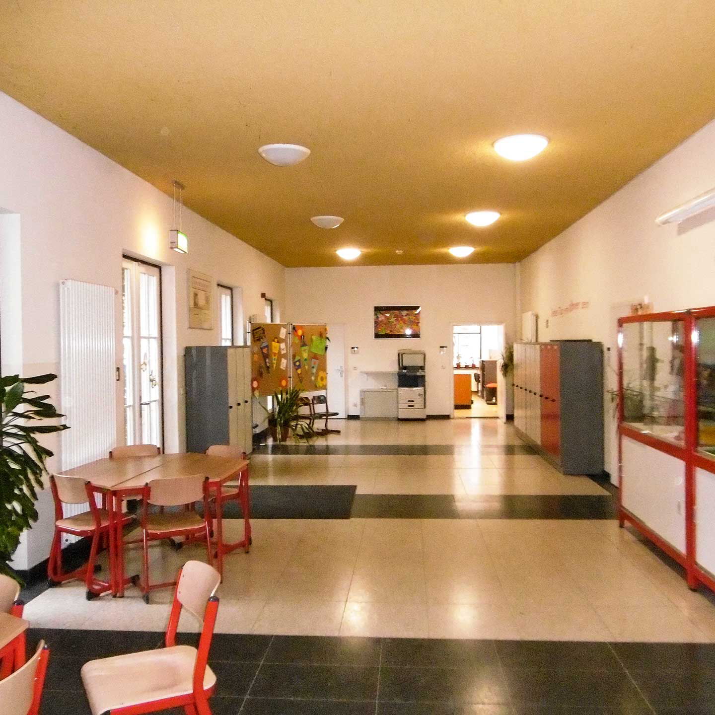 Der Foyerbereich der Grundschule mit einem langen Flur, mehreren Tischen, Schließfächern und zwei Vitrinen.