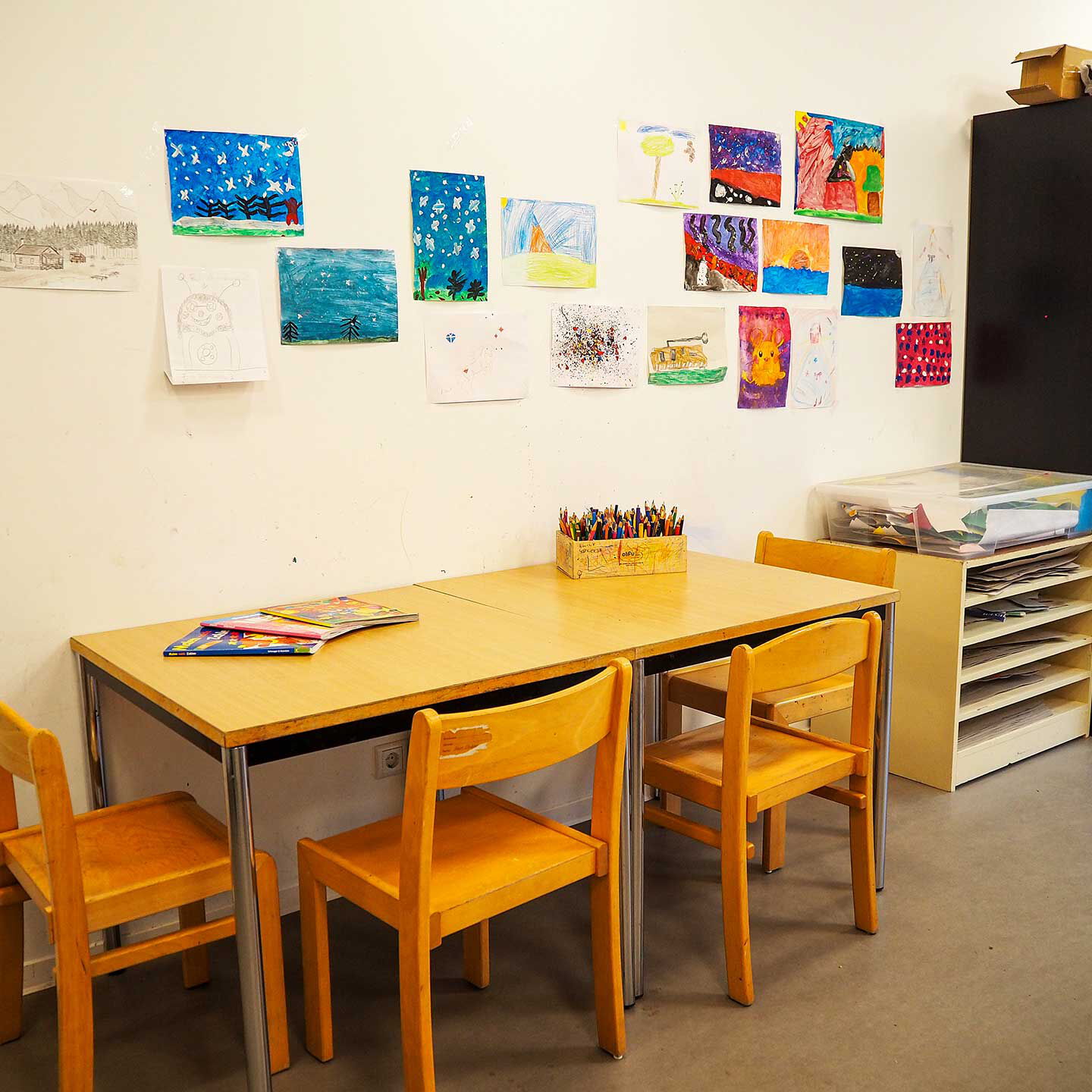 An der Wand stehen zwei Tische mit 4 Stühlen auf denen Buntstifte in einer Schachtel stehen. An der Wand hängen viele selbst gemalte Bilder.