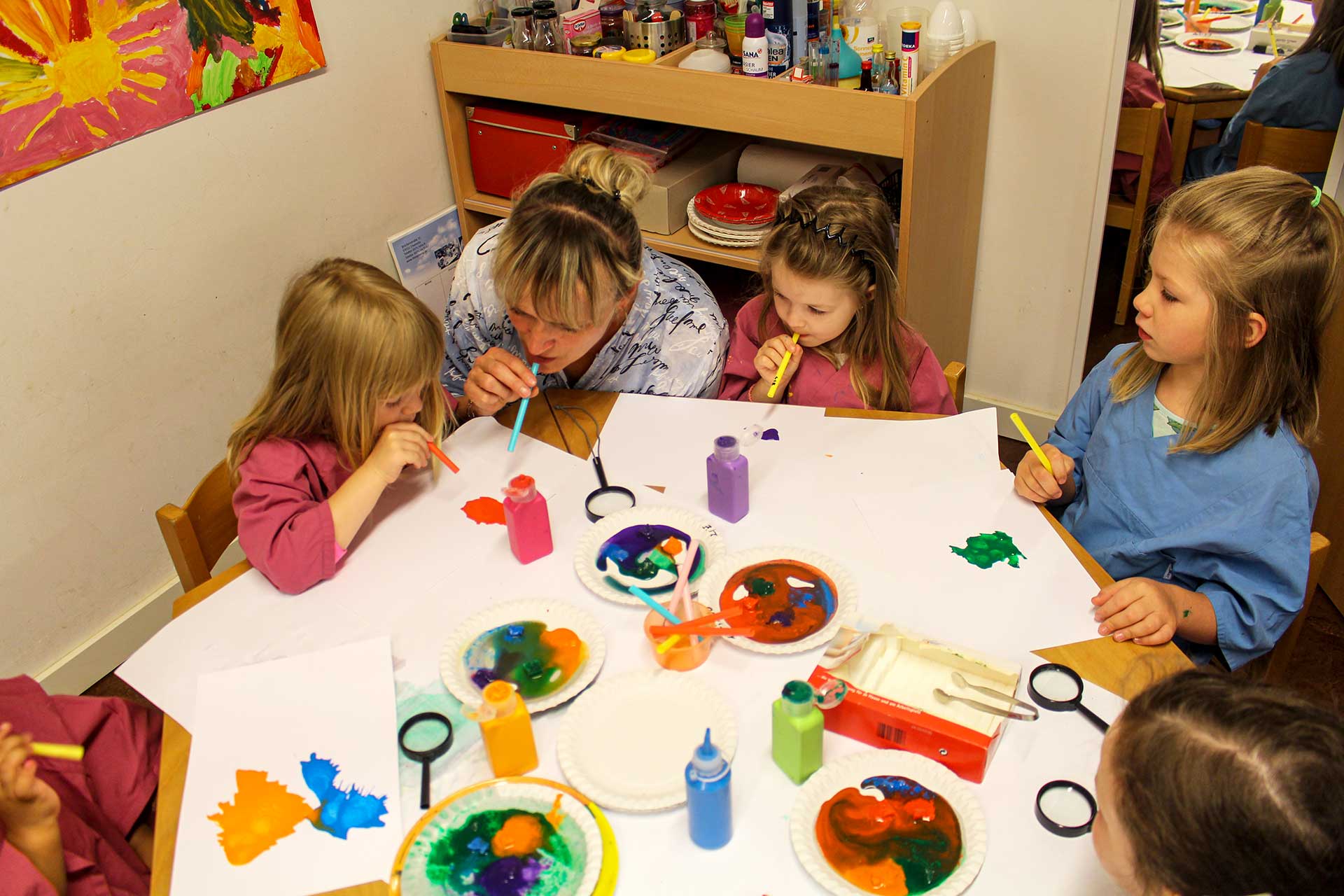 An einem Tisch sitzen fünf Kinder mit einer Erzieherin und pusten mithilfe eines Strohhalms Wasserfarben auf weiße Blätter.